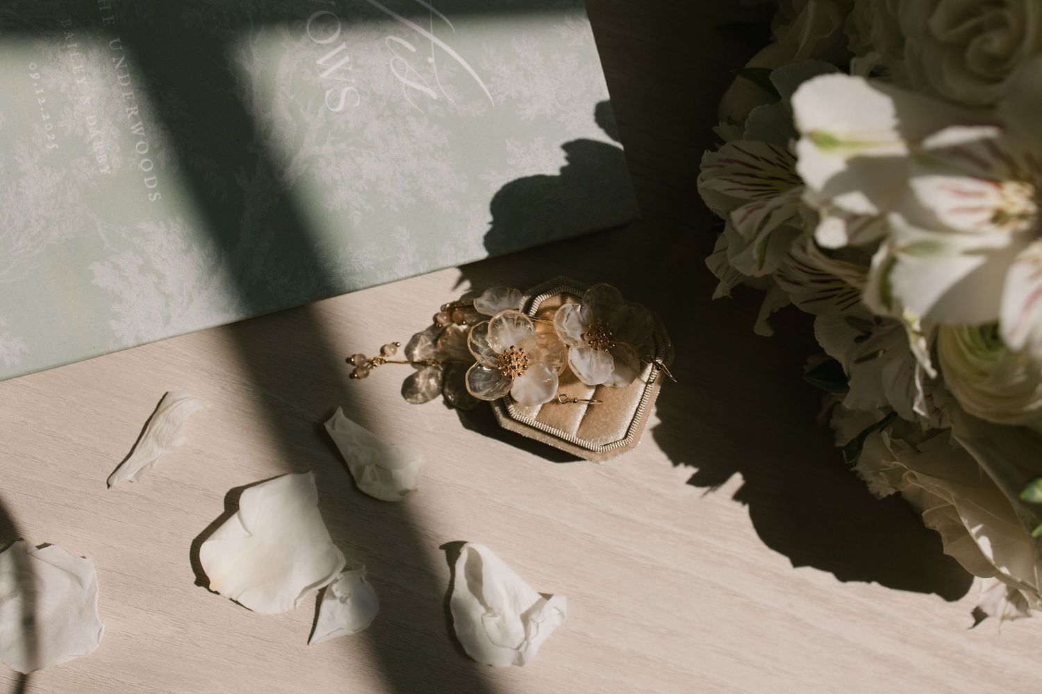 Gold charm bracelet and ring box with rose petals on sunlit surface beside white flowers.