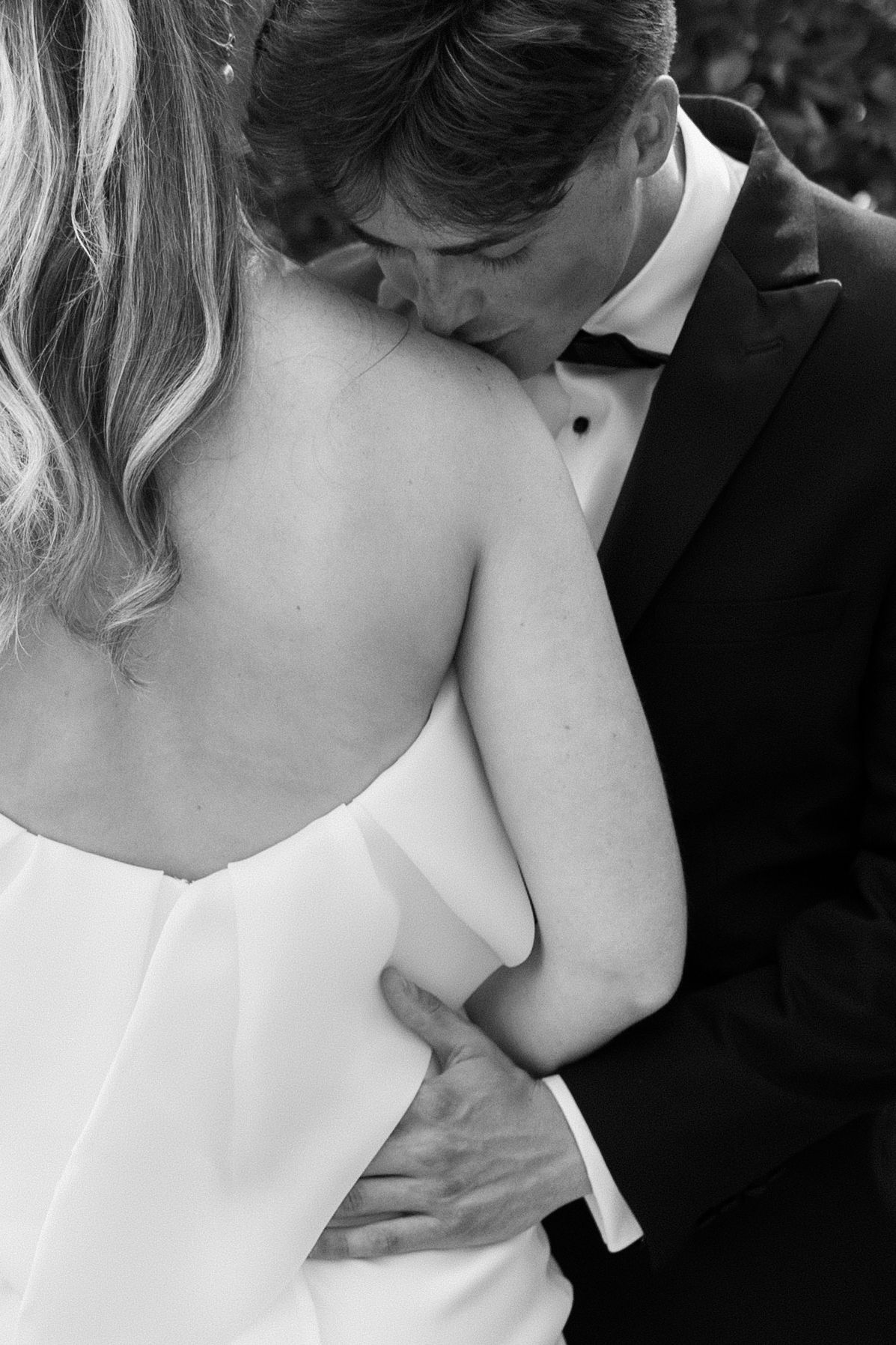 Black and white close-up of groom in tuxedo embracing bride in strapless wedding gown from behind.