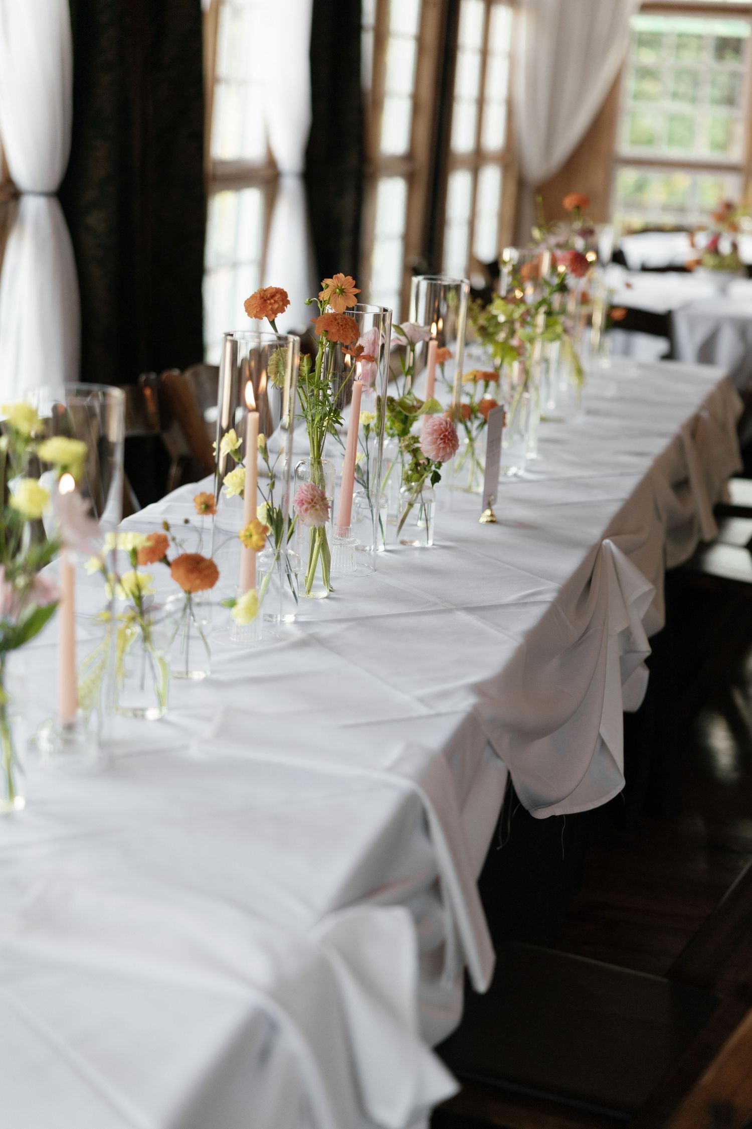Elegant wedding reception table with white linen, tall glass vases holding colorful spring flowers and candles.