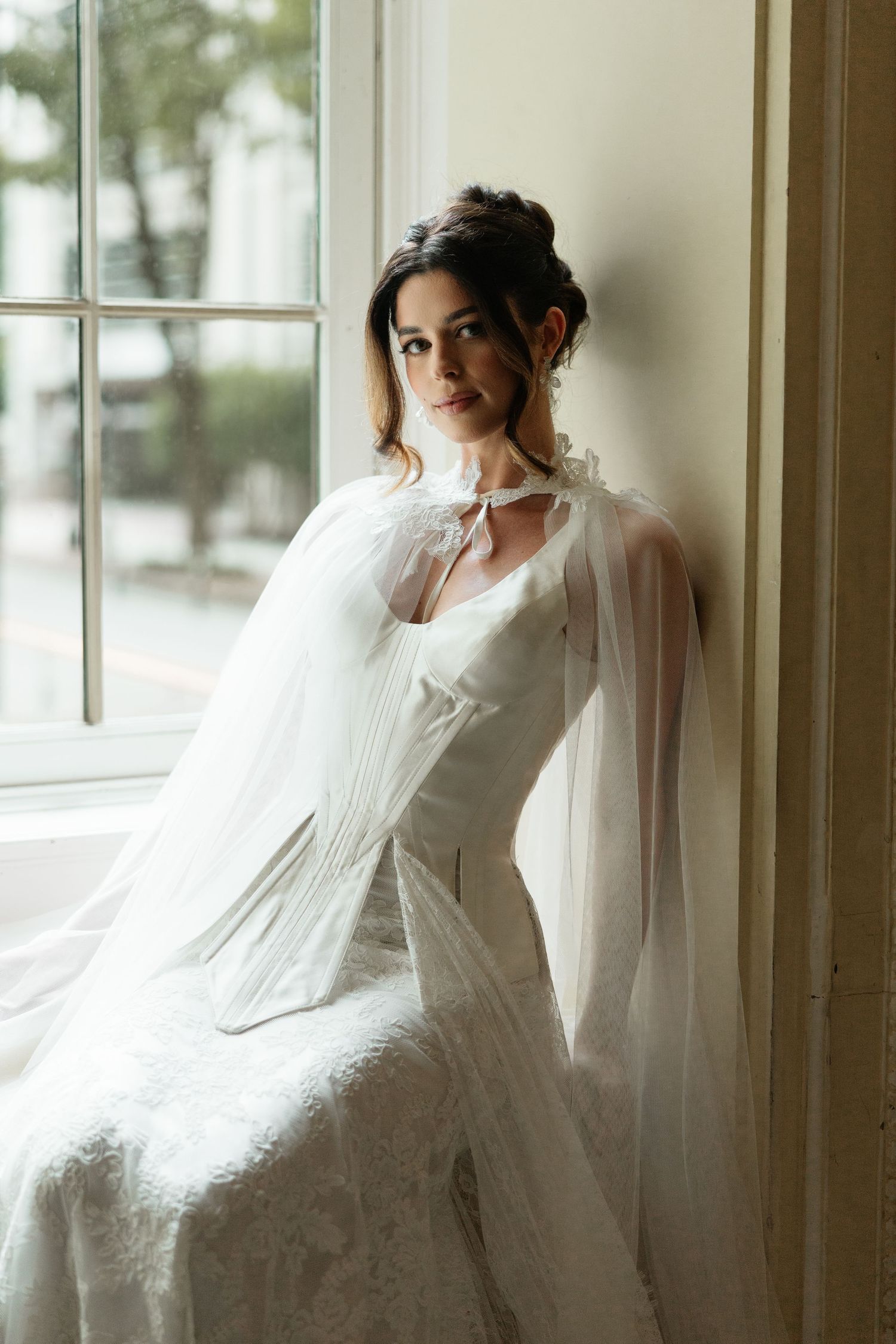 Bride in white lace gown and sheer cape sits by window, soft natural light highlighting her elegant updo.