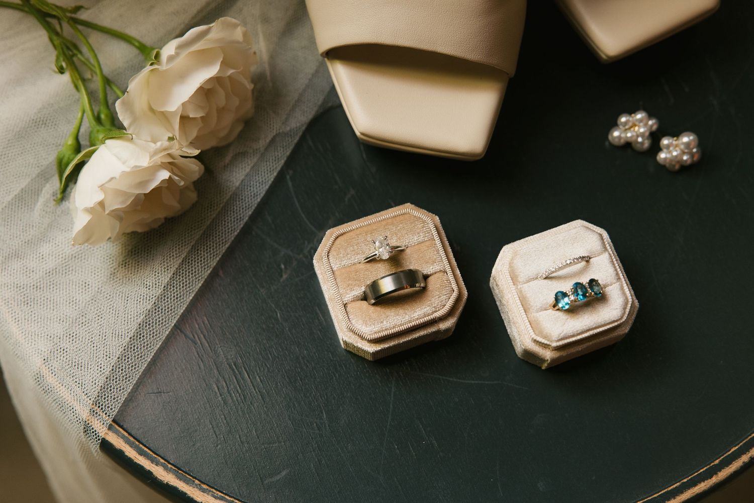 Close-up of two velvet ring boxes with wedding rings, pearl earrings, and white roses on a dark green surface.