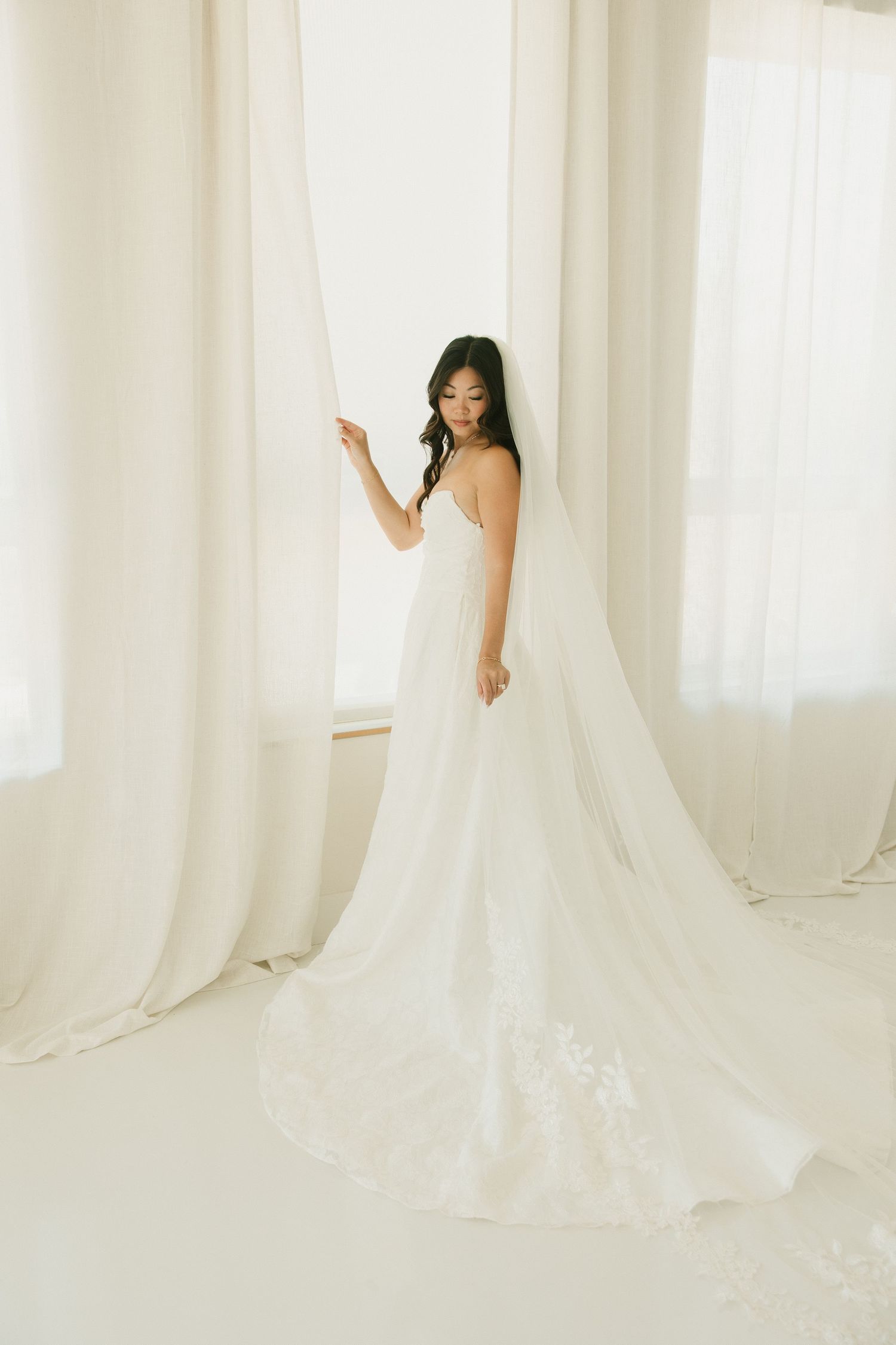 An elegant bride in a fitted lace wedding gown with a long veil stands by sheer white curtains.