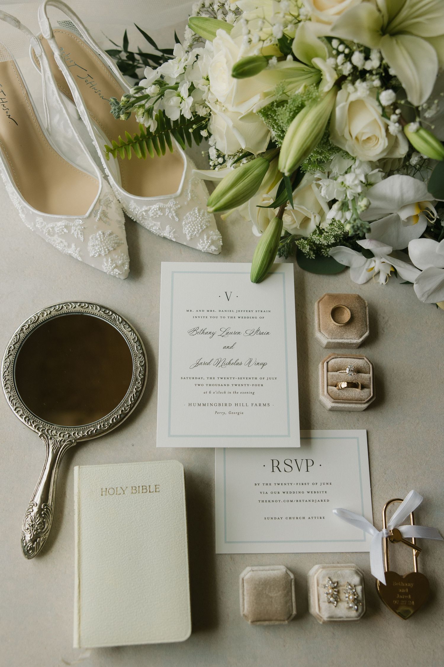 Elegant wedding flat lay with invitation, RSVP card, rings, shoes, bouquet, and Holy Bible.