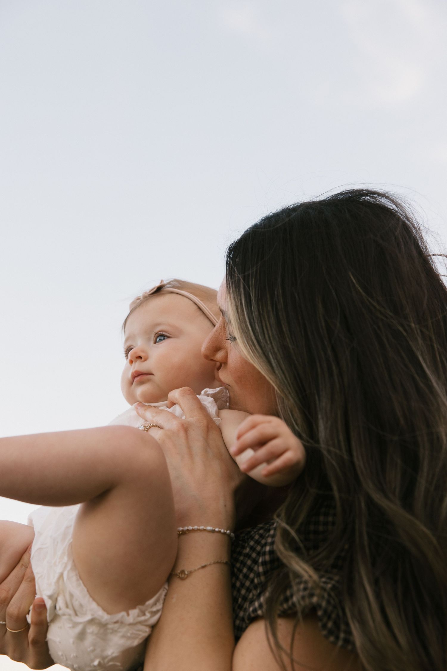 A dark-haired mother gently kisses her wide-eyed baby girl against a soft bright sky background.