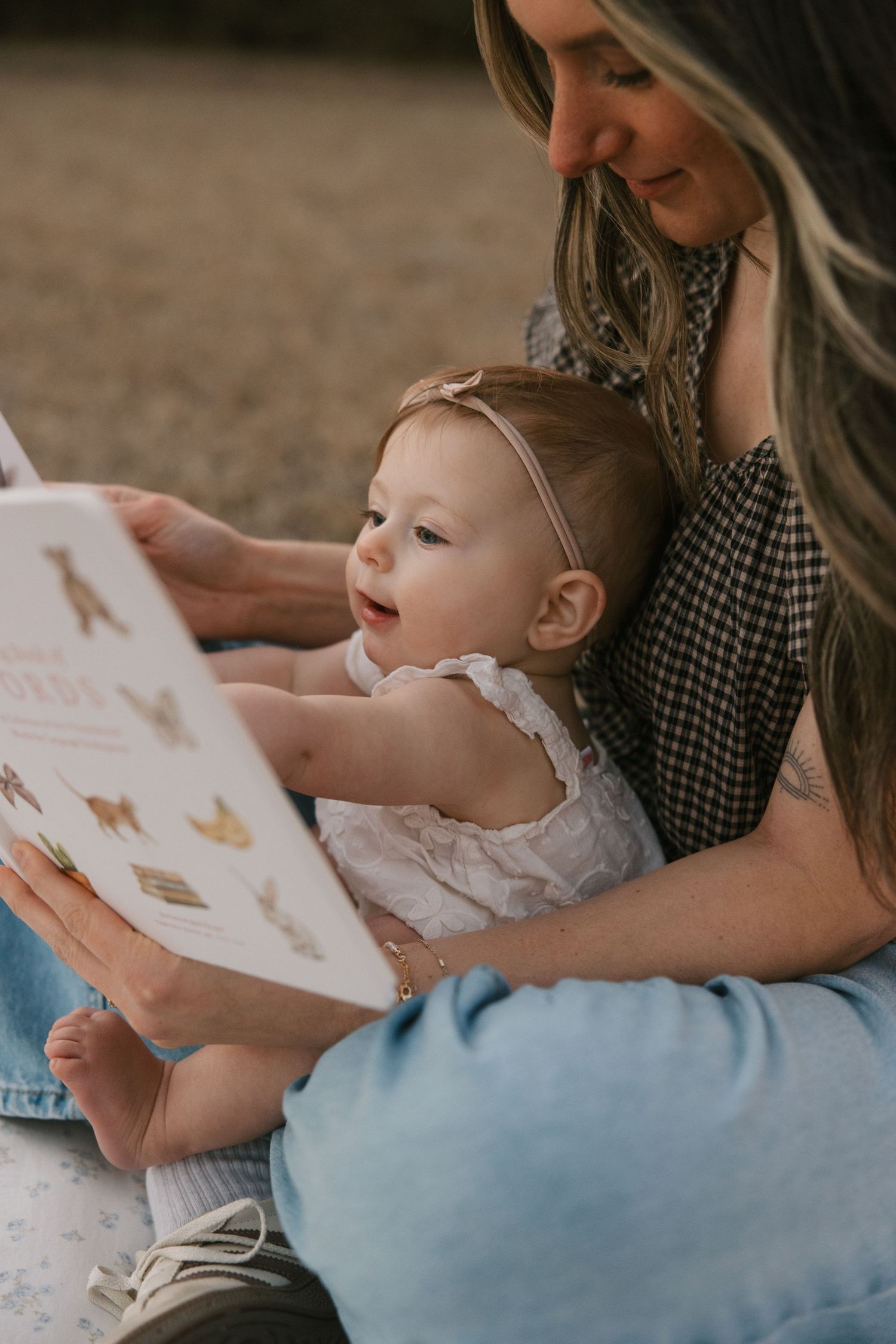 A mother with blonde highlights reads a colorful picture book to her curious baby girl outdoors.