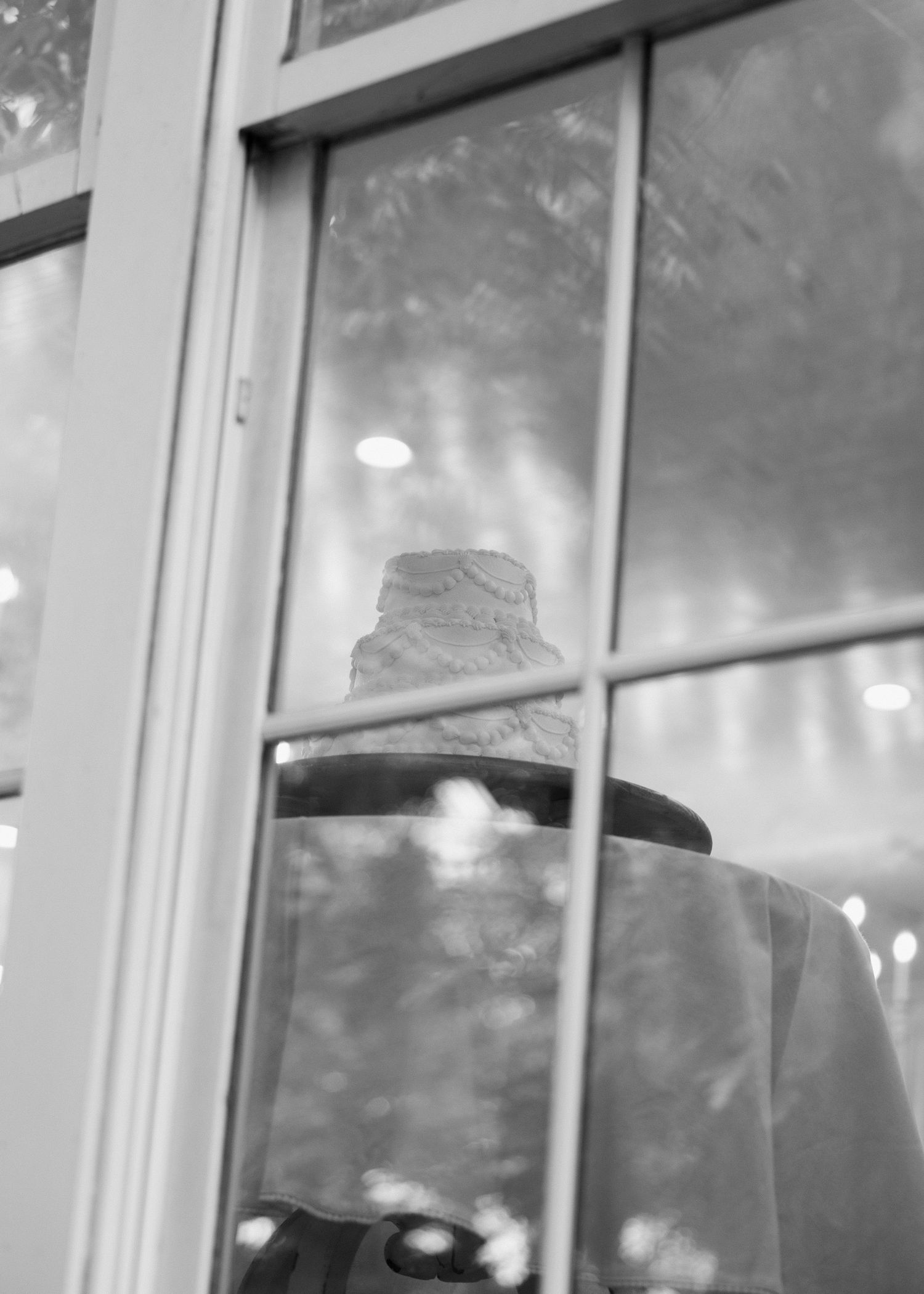 Black and white photo of a tiered wedding cake on a draped table, viewed through a glass door with bokeh lights.