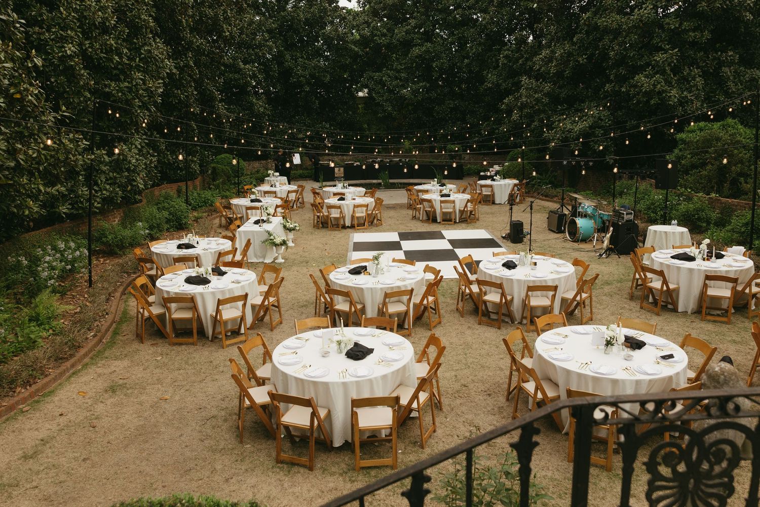 Outdoor wedding reception setup with round tables, wooden chairs, string lights, and a checkered dance floor in a garden.
