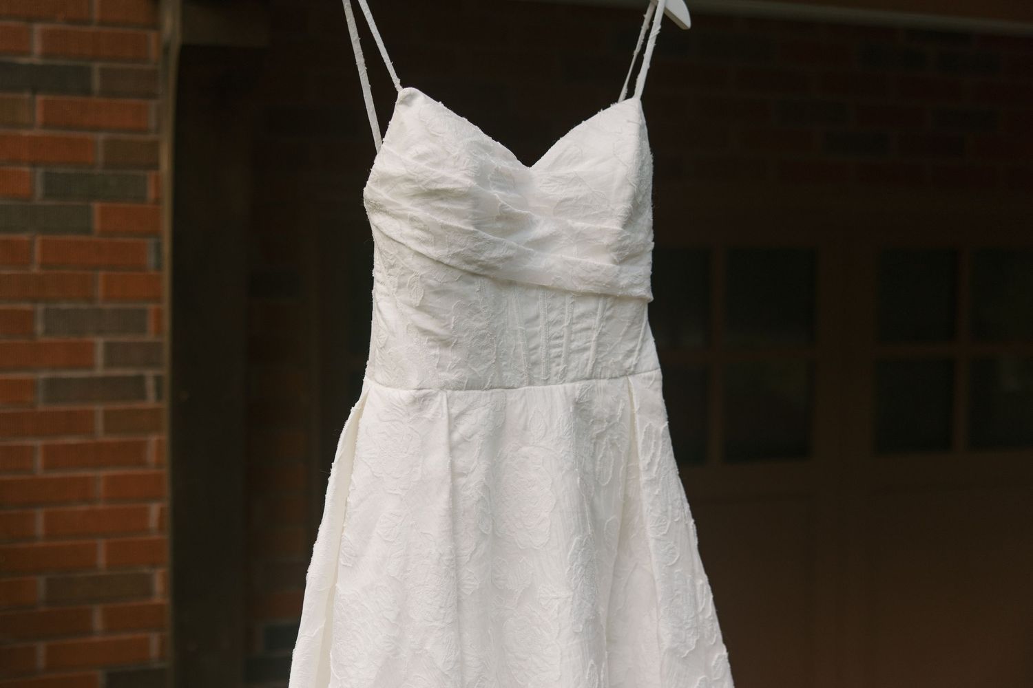 White spaghetti strap wedding dress with sweetheart neckline hanging outdoors near brick wall.