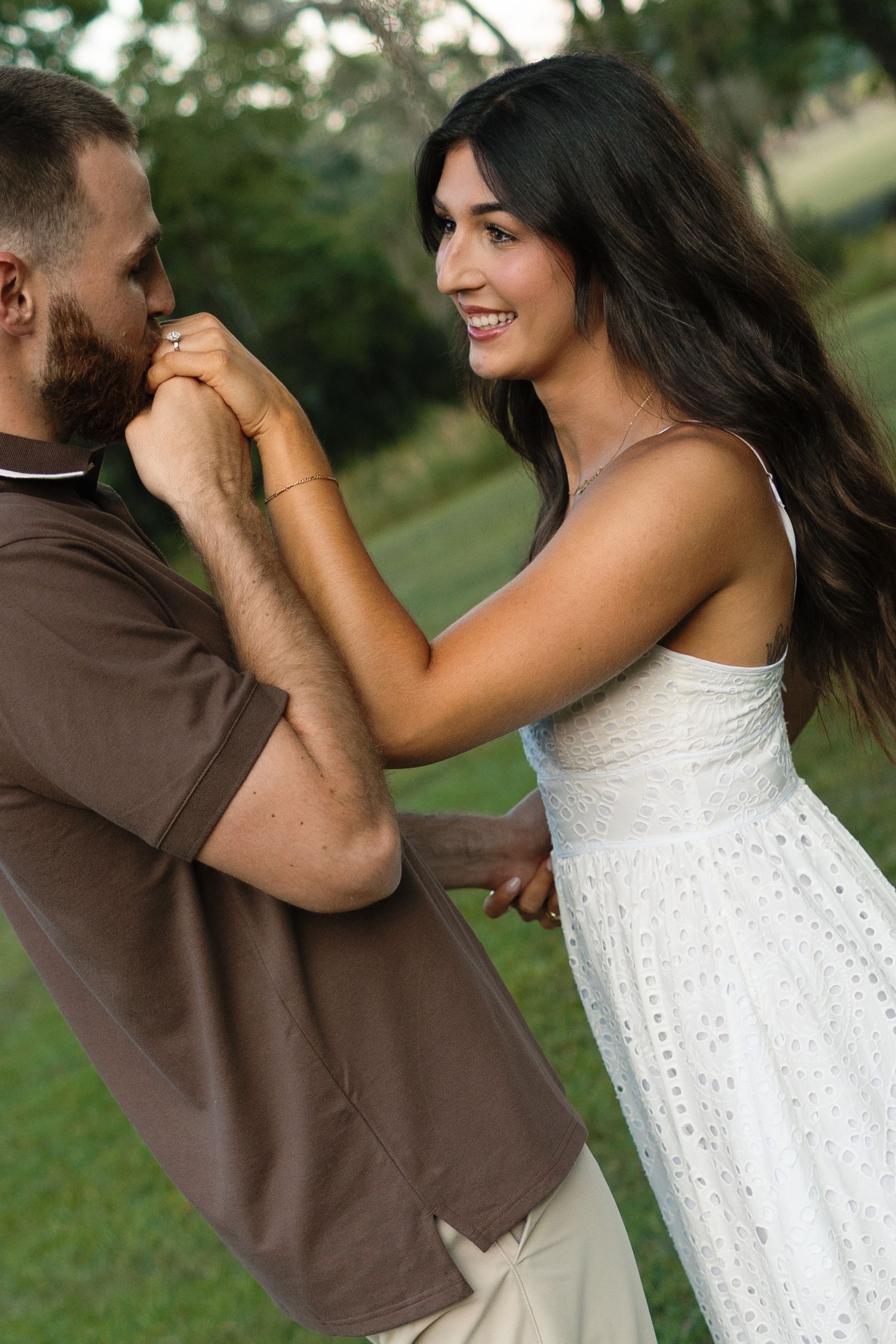 Romantic couple dancing outdoors, woman in white dress smiling as man kisses her hand in a lush green park.