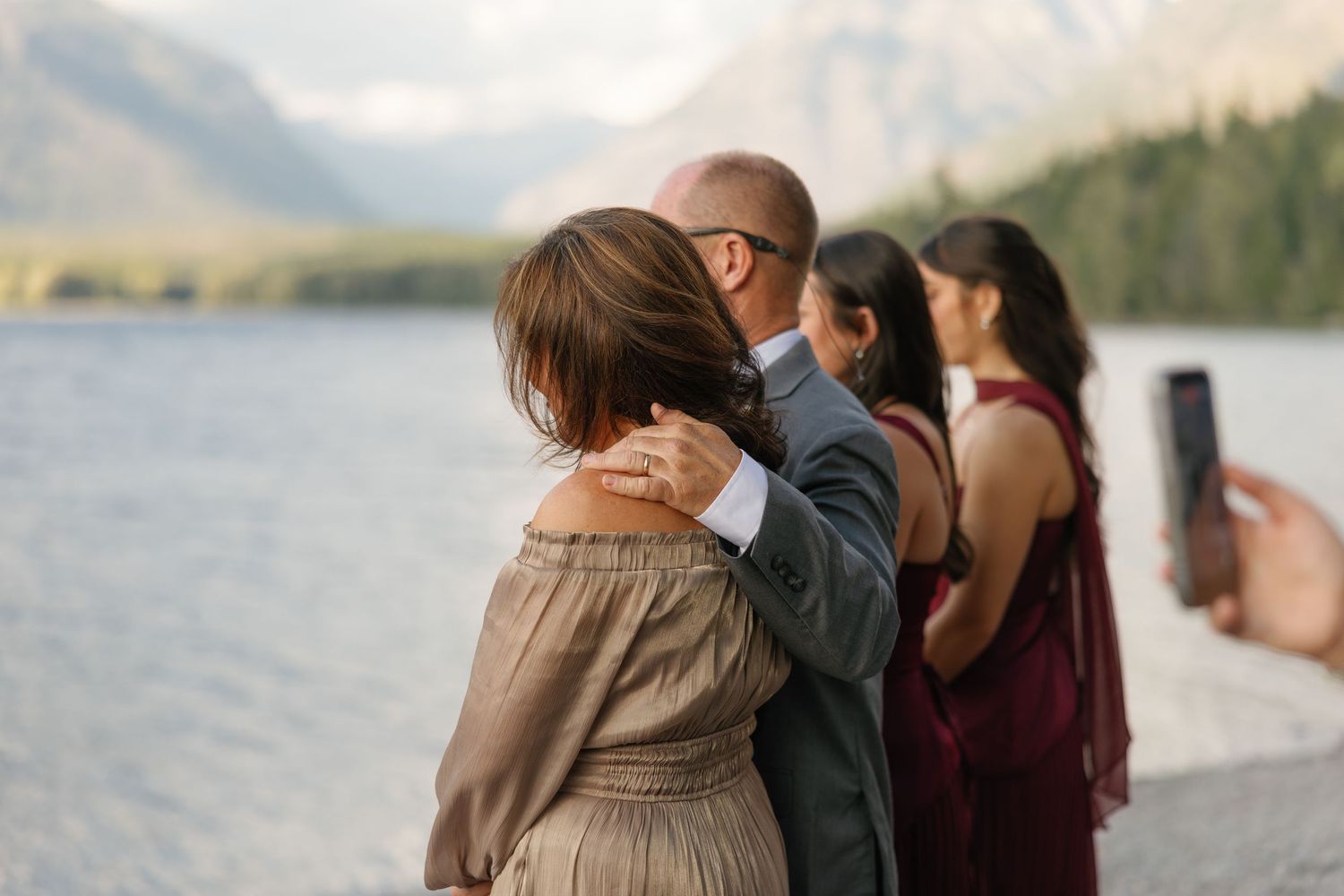 Couple embracing by a mountain lake during an outdoor wedding ceremony with guests nearby.