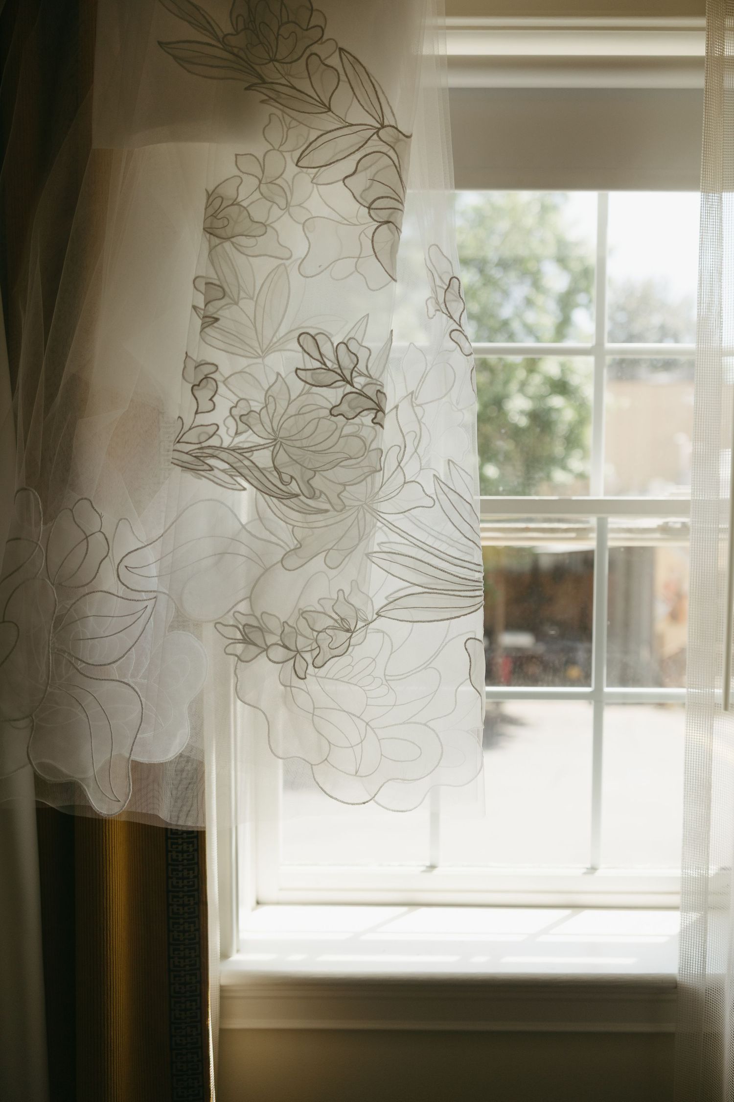 White floral embroidered wedding dress hanging near a sunlit window with soft natural light.