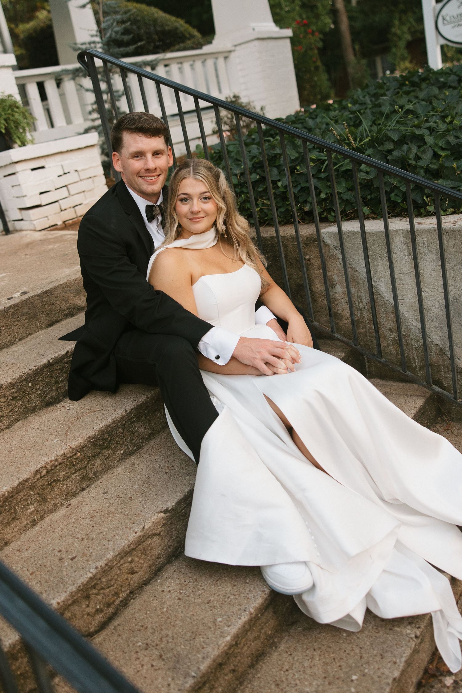 Bride and groom sitting on stone steps outdoors, she in a white gown, he in a black tuxedo, smiling warmly.