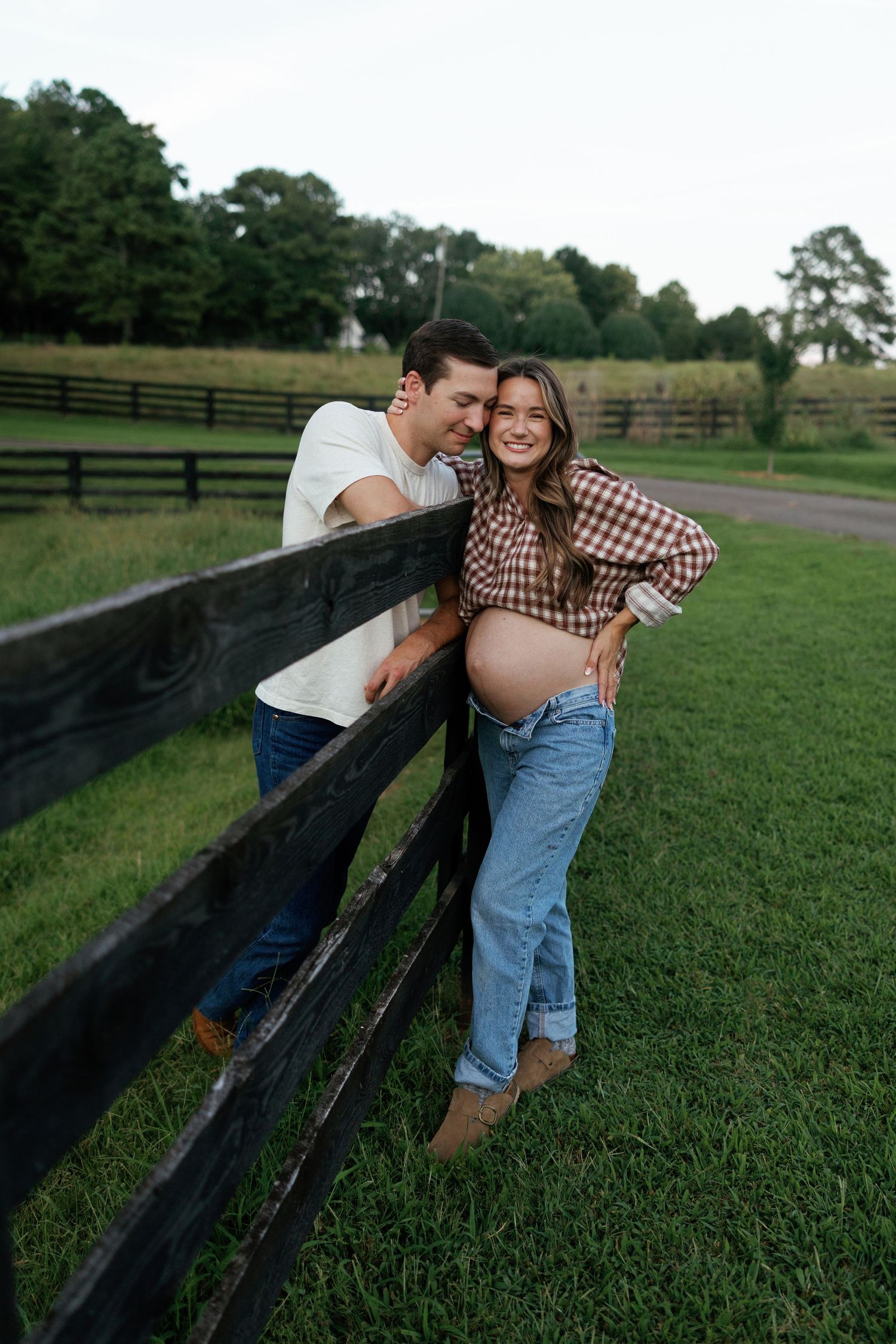 Couple posing by a wooden farm fence, woman showing pregnant belly in checkered shirt and jeans, green pasture background.
