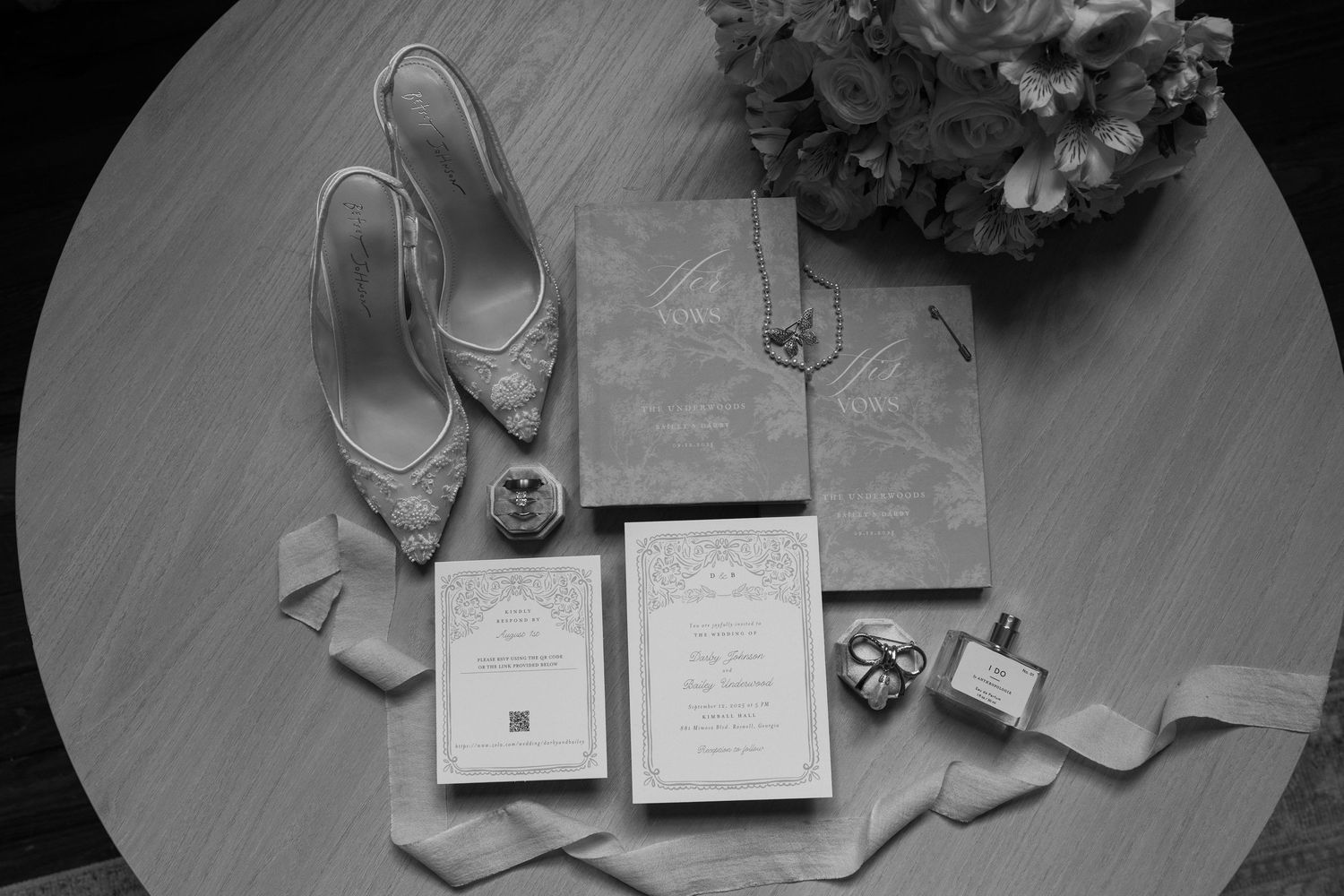 Black and white wedding flat lay with heels, invitations, rings, perfume, and floral bouquet on round table.