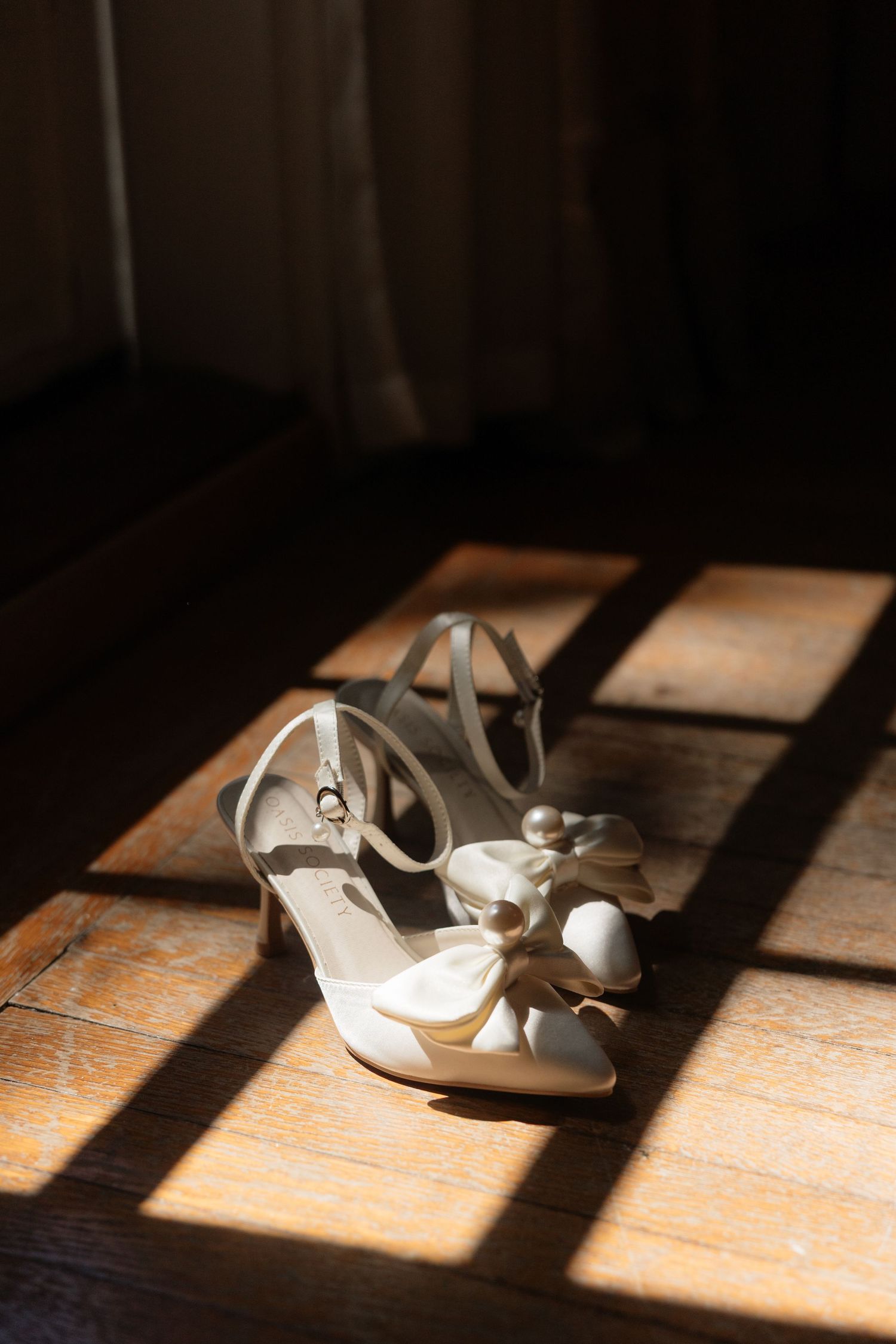 Elegant white bridal heels with floral bow detail resting on wooden floor in warm sunlight.