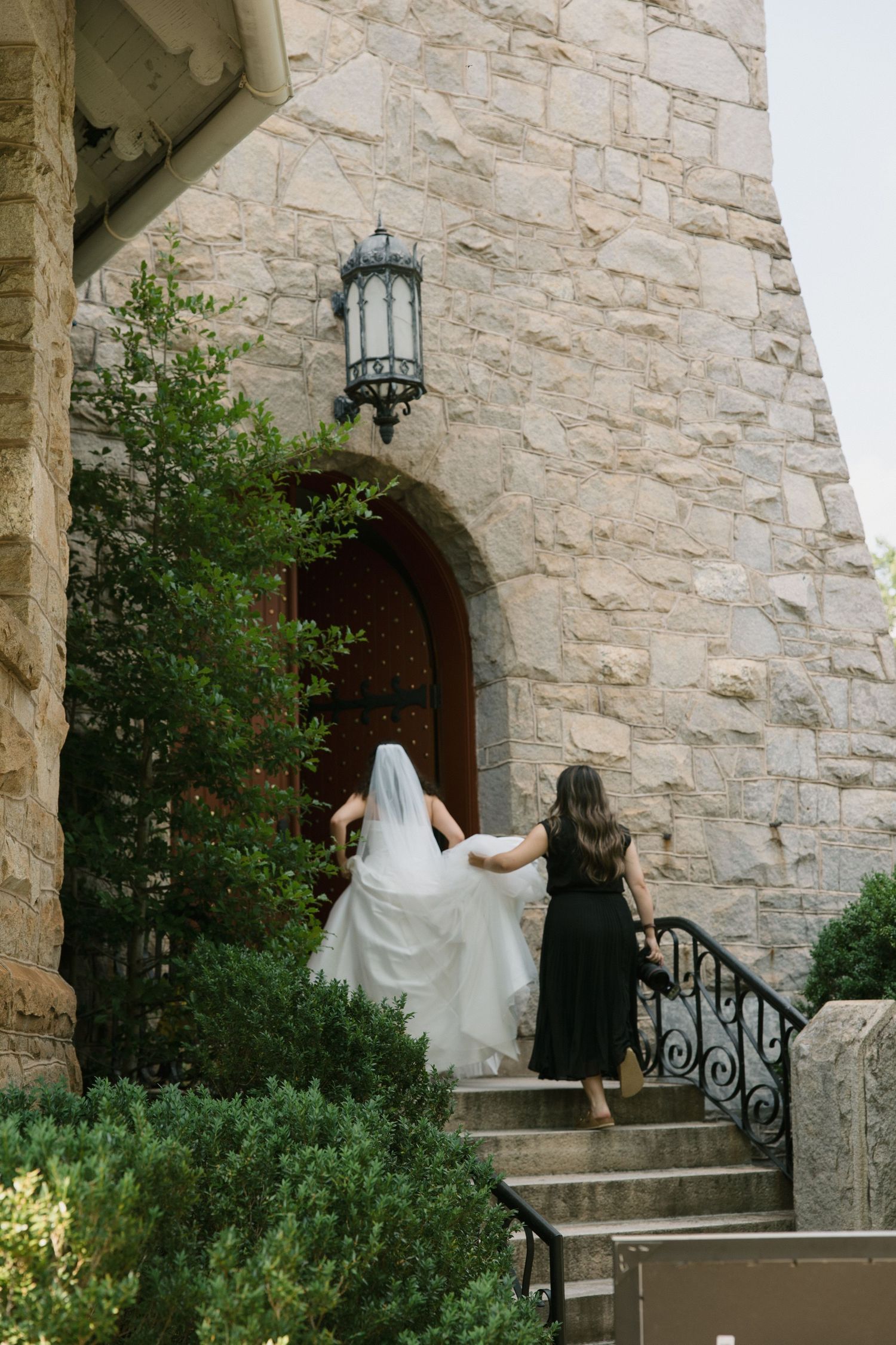 Bride in white gown and veil walks up stone church steps with bridesmaid in black dress holding her train.