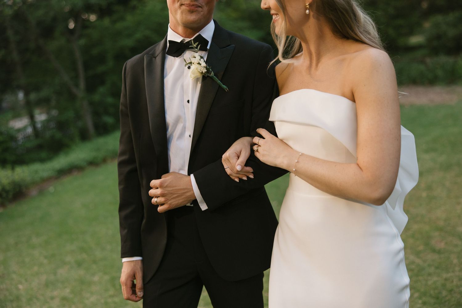 Bride in strapless white gown walks arm in arm with groom in black tuxedo and bow tie on lush green lawn.