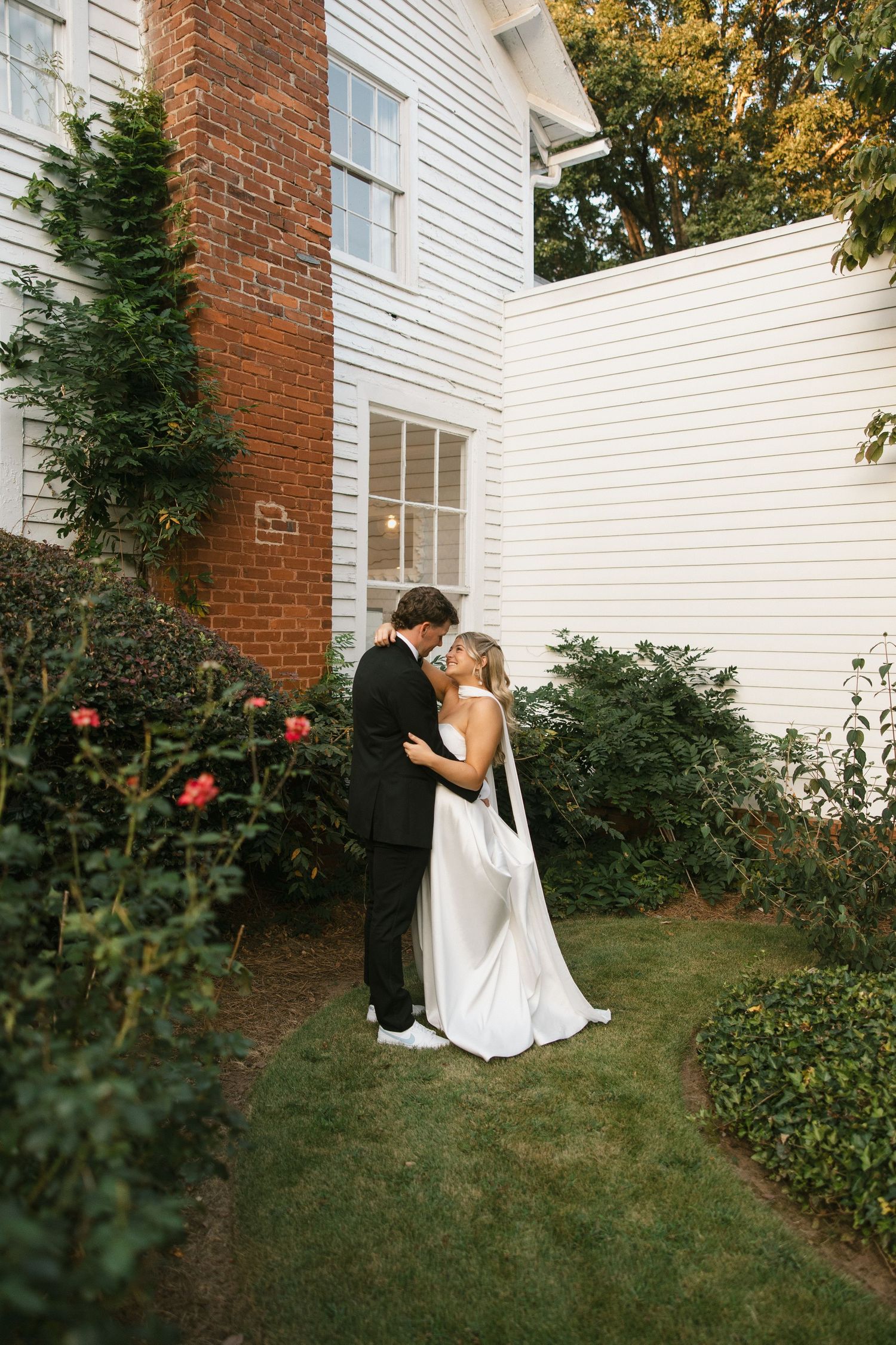 Bride and groom share a romantic kiss in a lush garden beside a white colonial home with brick chimney.
