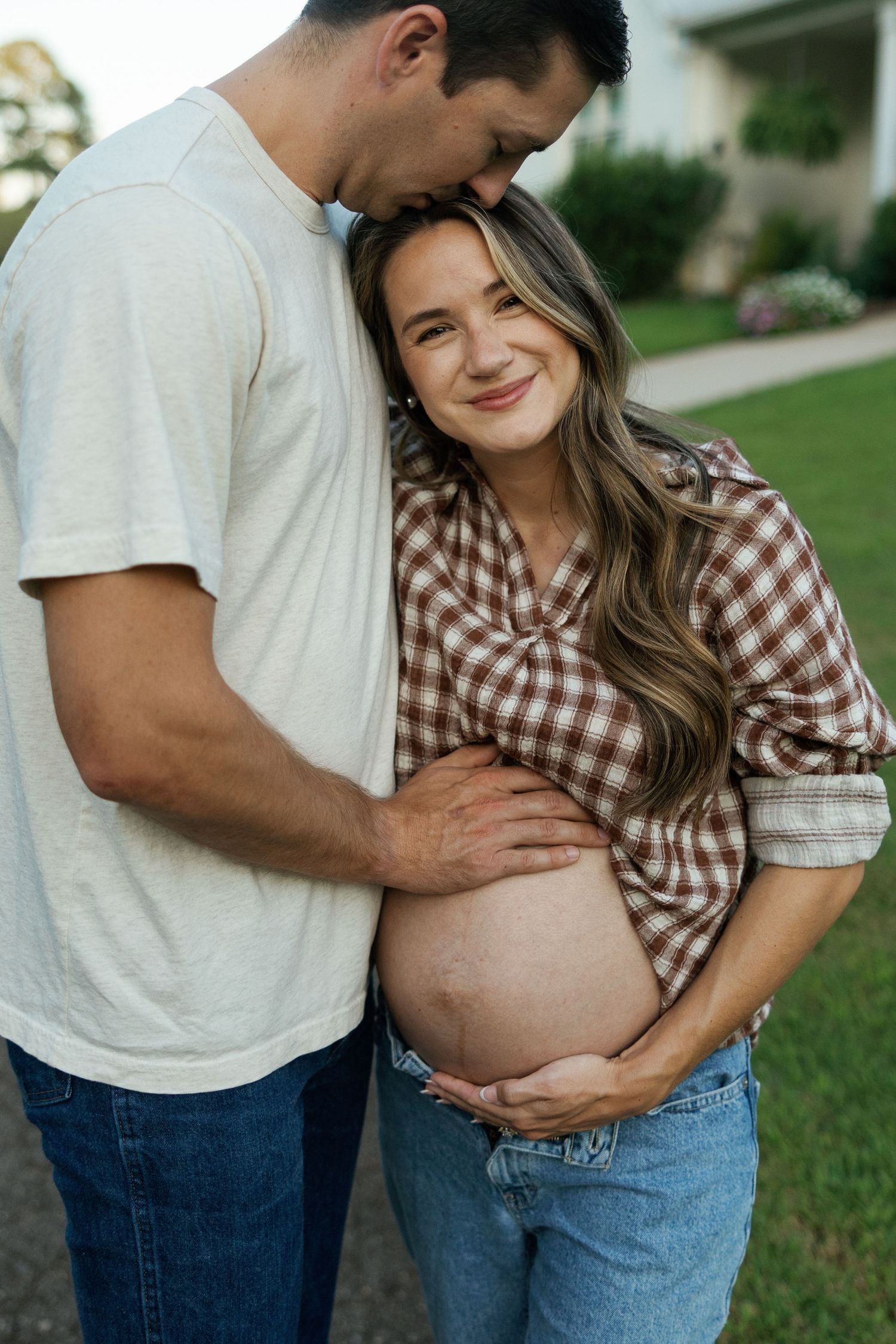 A smiling pregnant woman in plaid shirt and jeans embraced by her partner outdoors during a maternity photo session.