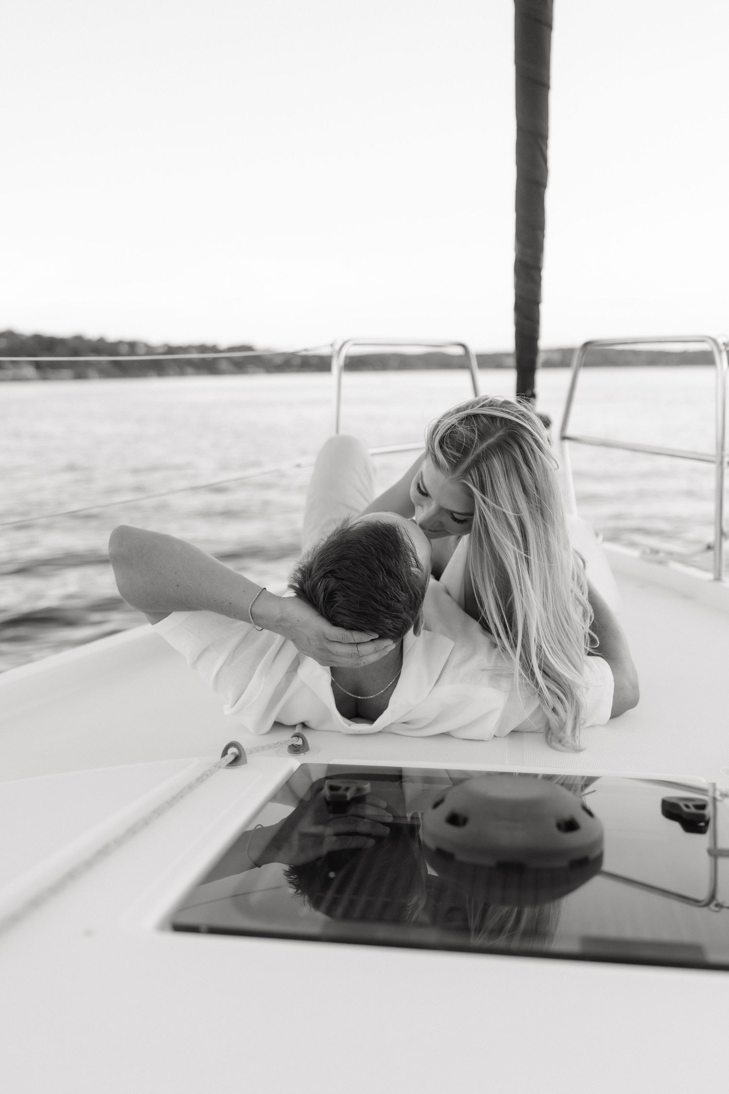 Black and white photo of a romantic couple cuddling on the bow of a sailboat on calm open water.