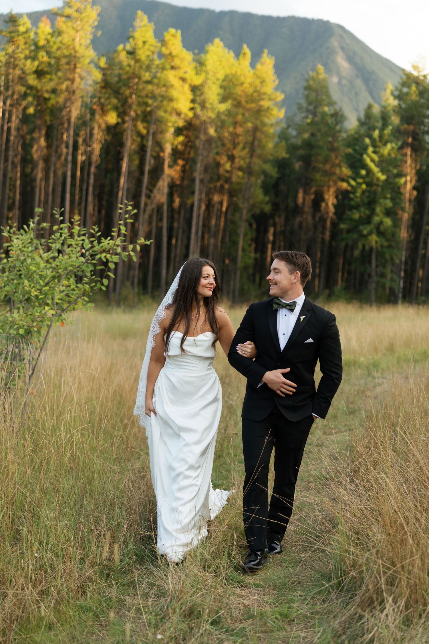 Bride and groom walking through mountain meadow, she in white gown with veil, he in black tuxedo.