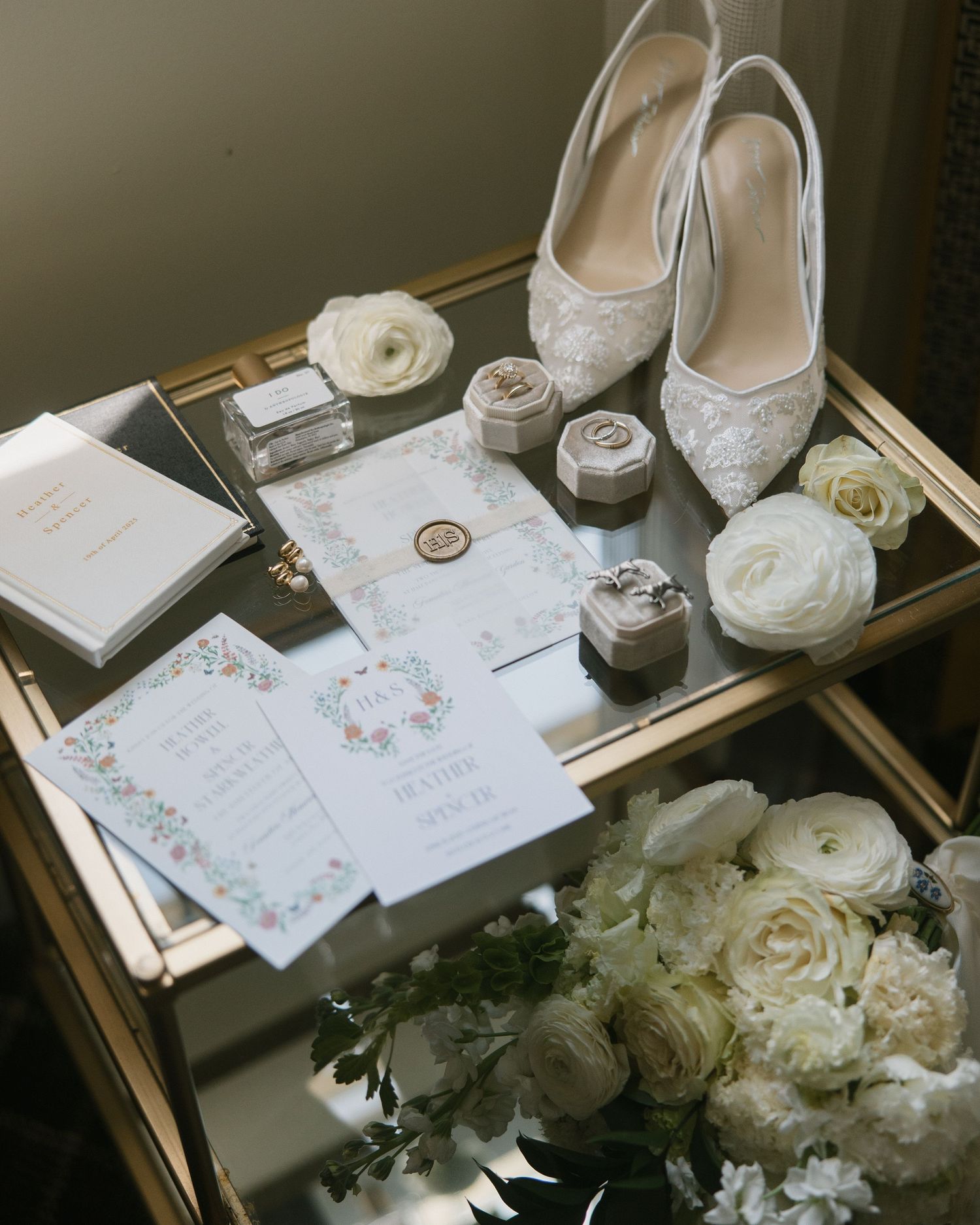 Elegant wedding flat lay with lace heels, floral invitations, ring boxes, white roses, and perfume on a gold mirror tray.