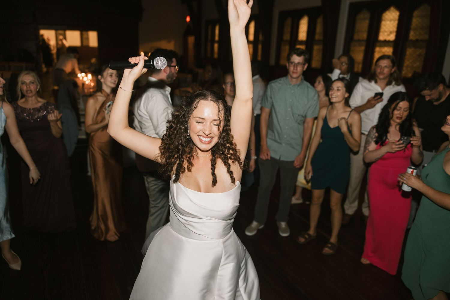 Bride in white gown dancing joyfully with arm raised at wedding reception surrounded by guests.