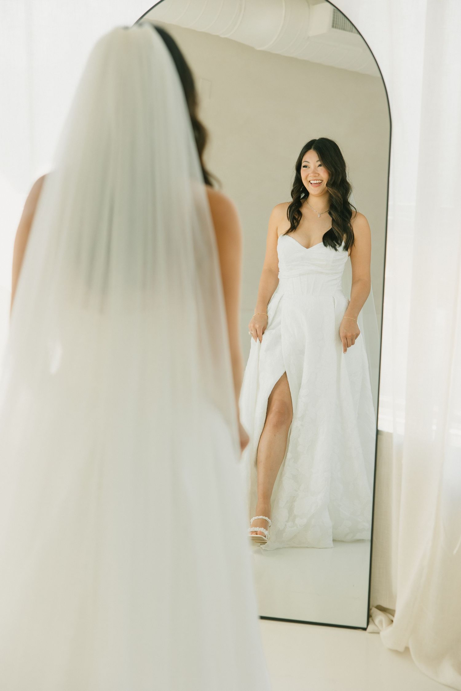 A smiling bride in a white slit wedding gown admires her reflection in a large arched mirror.