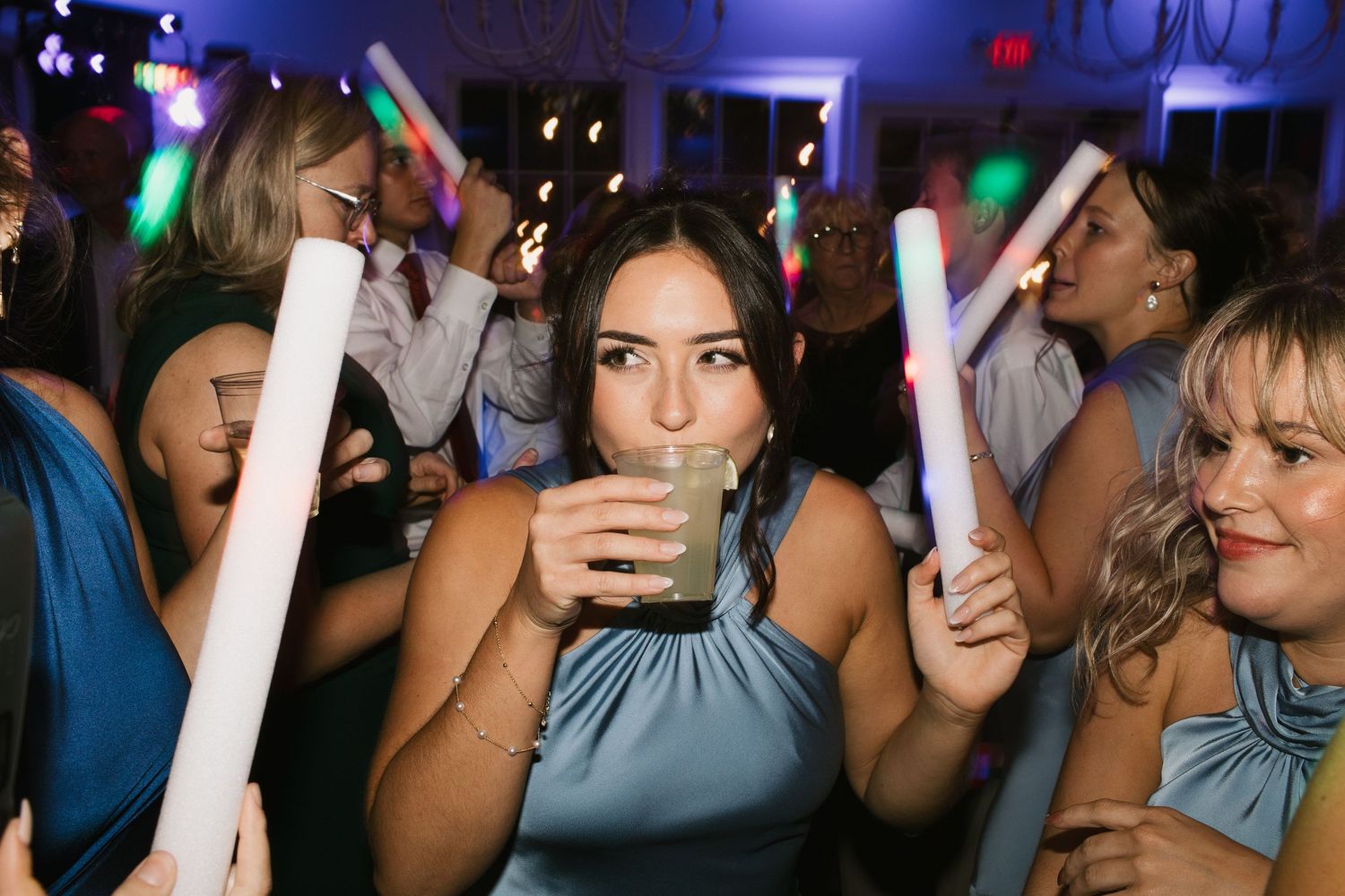 Bridesmaid in a blue dress holding a drink and glow stick on a colorful wedding dance floor.