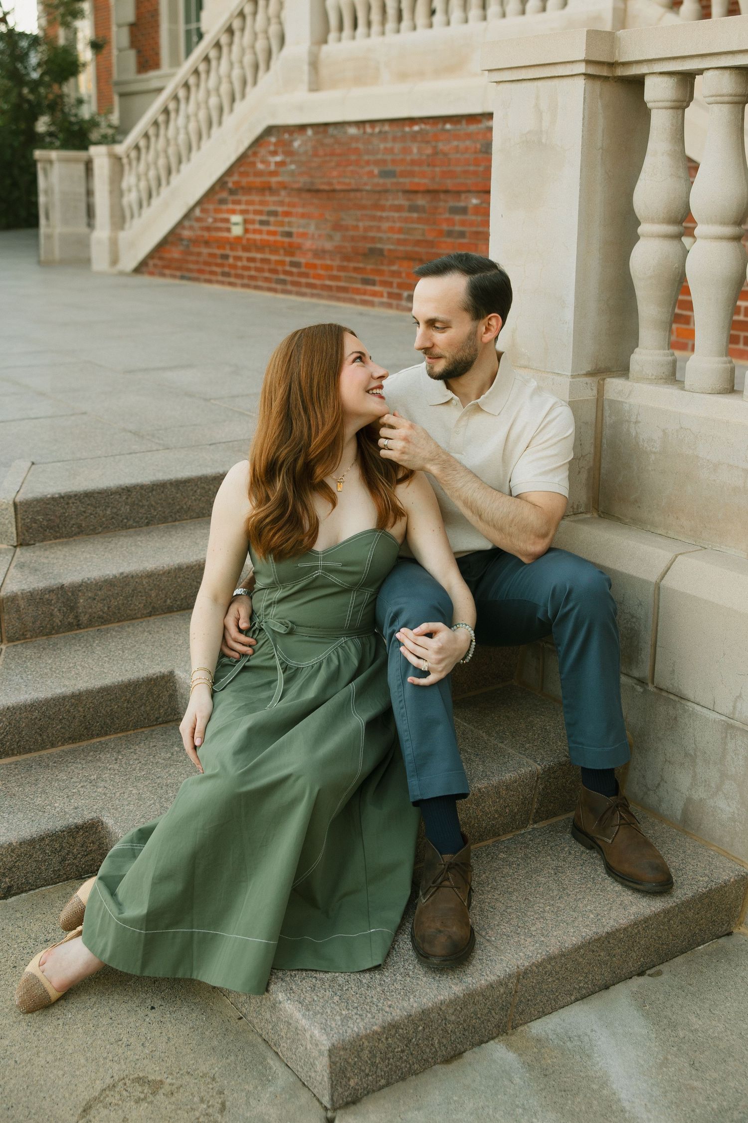 A couple sits on stone steps, man in cream polo and blue pants, woman in elegant green dress.