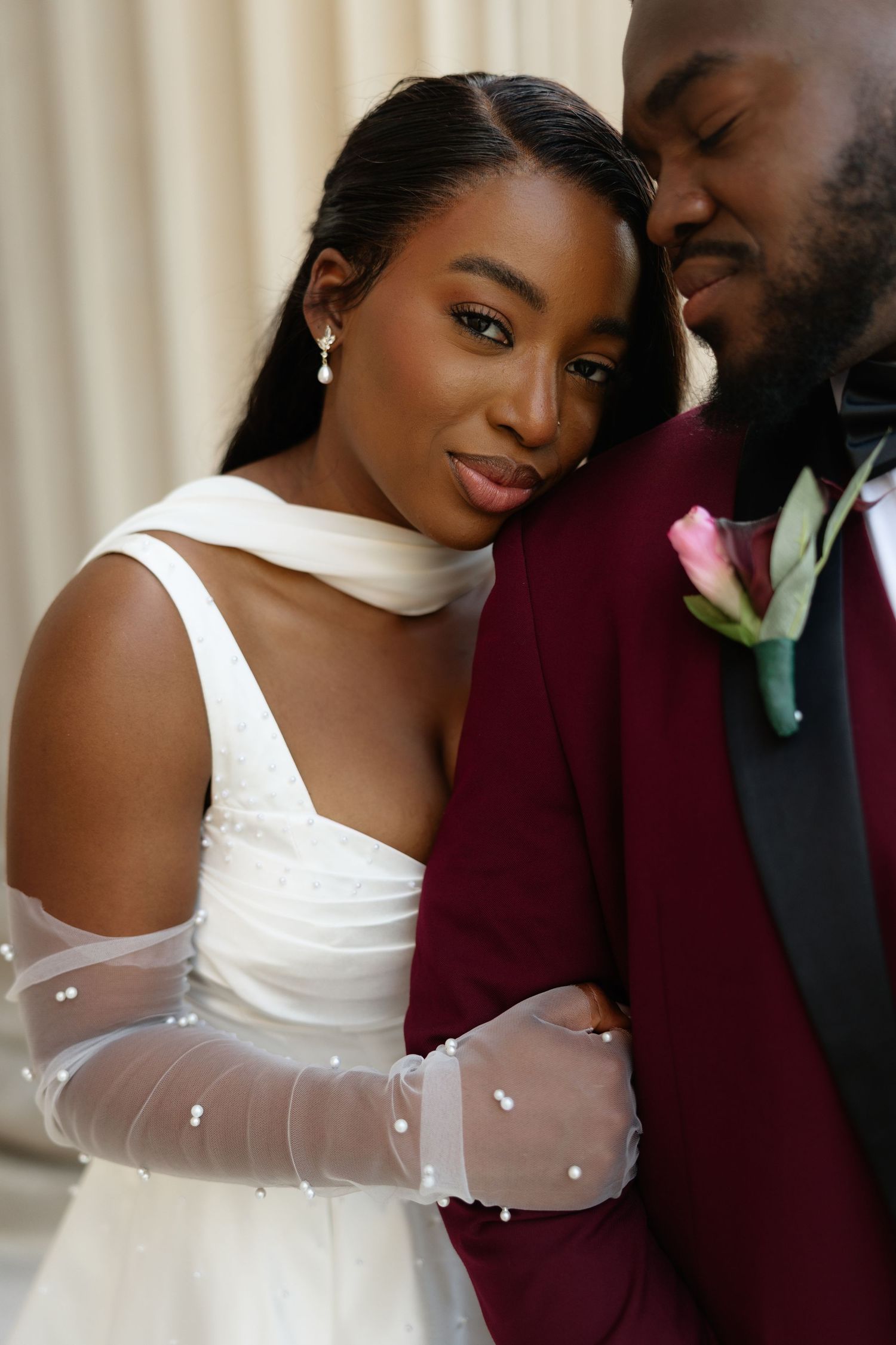 Elegant Black bride in white gown with sheer gloves leans on groom in burgundy suit with pink boutonniere.