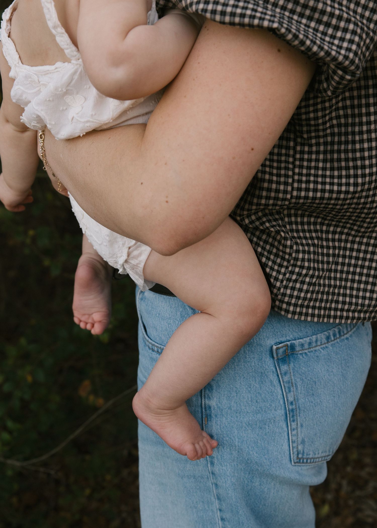 A parent in a plaid shirt and jeans cradles a tiny barefoot baby against their side outdoors.