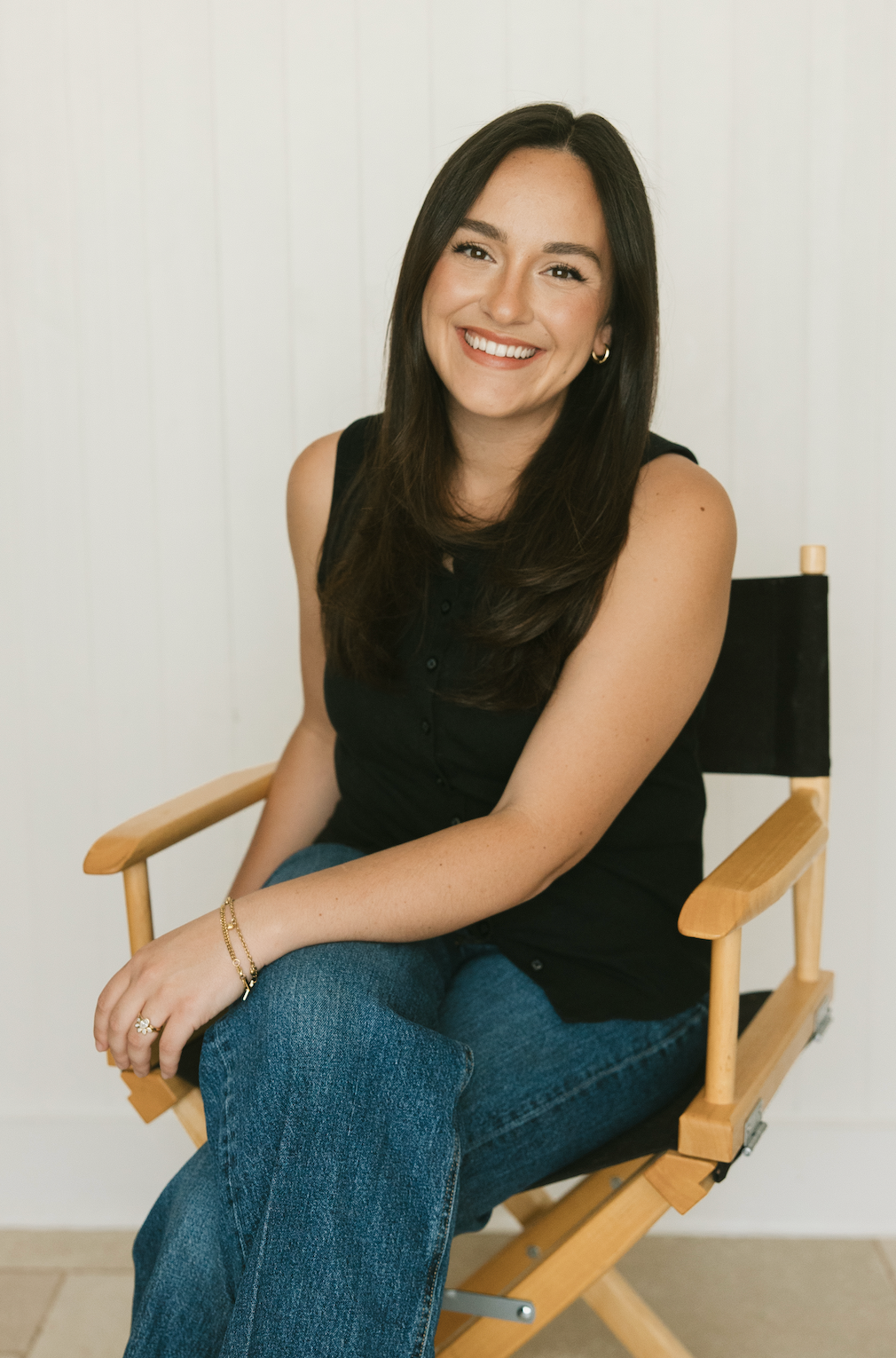 Smiling young woman with long dark hair sitting in a director's chair wearing a black top and jeans.