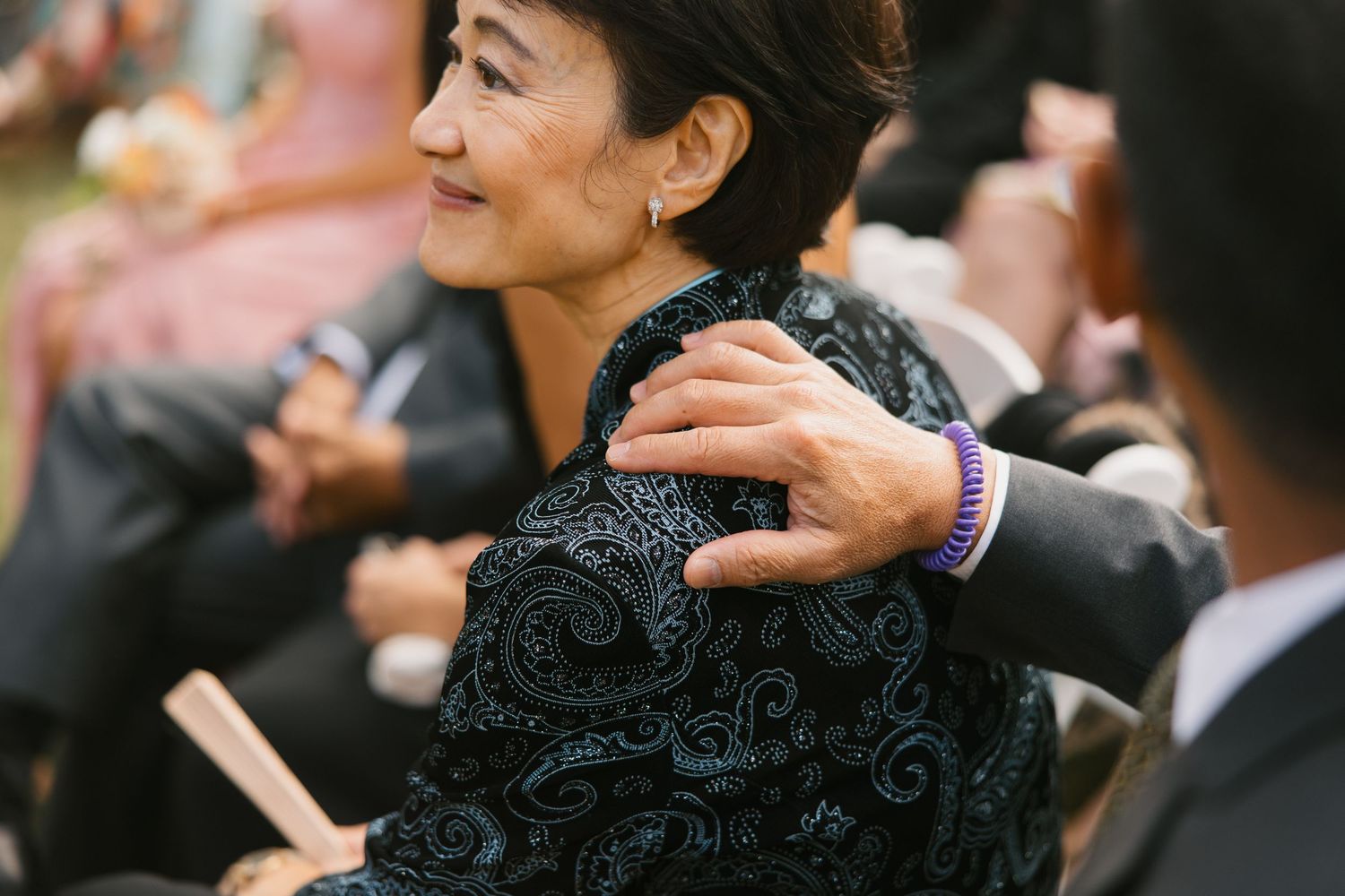 Wedding guest in floral dress smiles warmly as someone places a hand on her shoulder during ceremony.