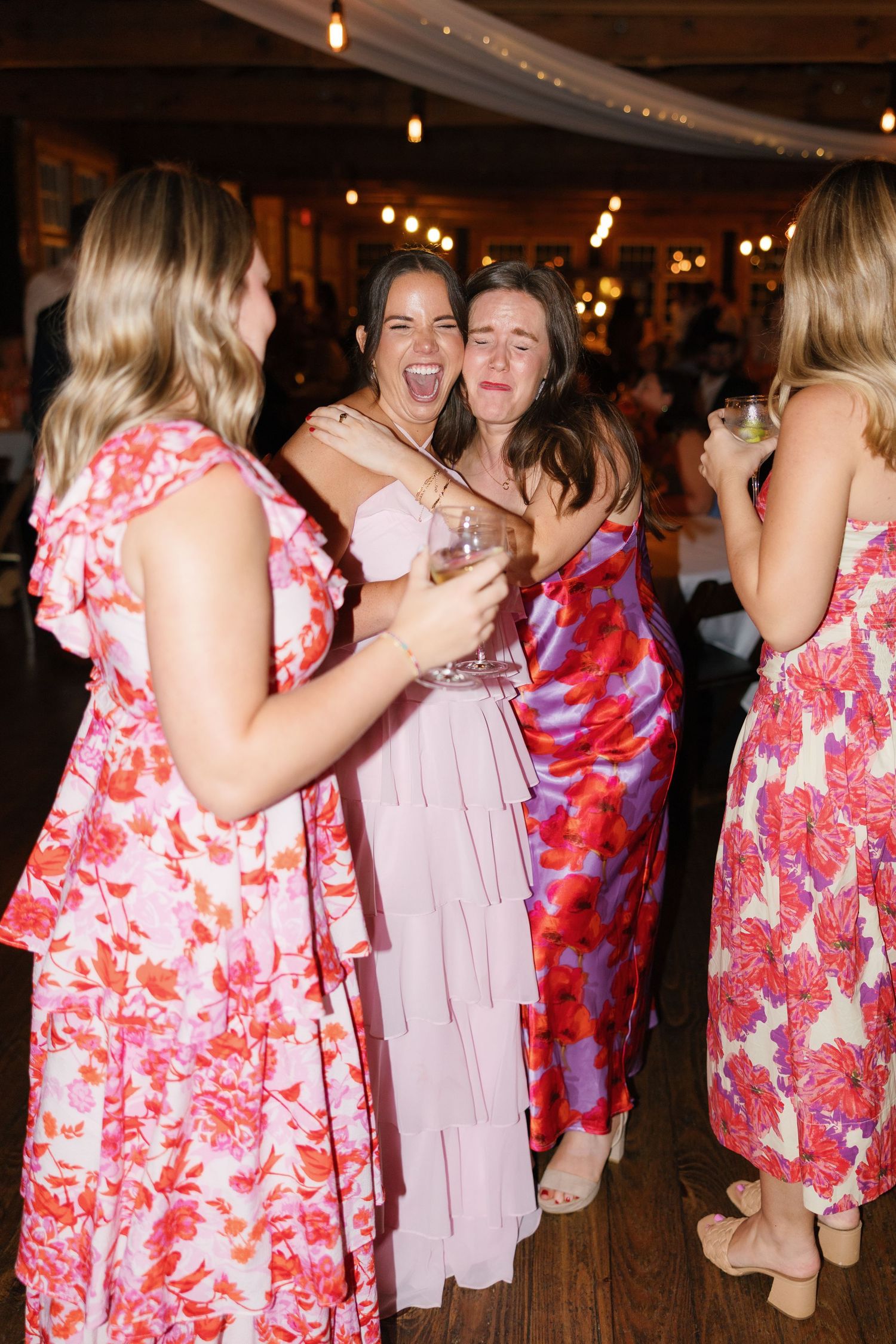 Three women in floral dresses laughing and dancing together at an indoor wedding reception celebration.