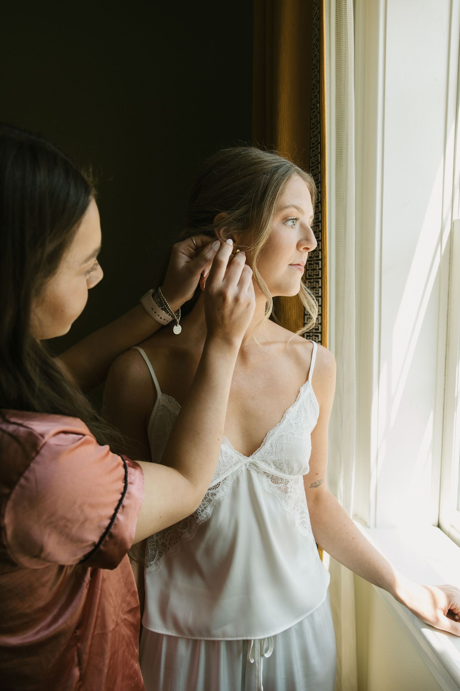 Bridesmaid in pink robe helping bride put on pearl earring by a sunlit window.
