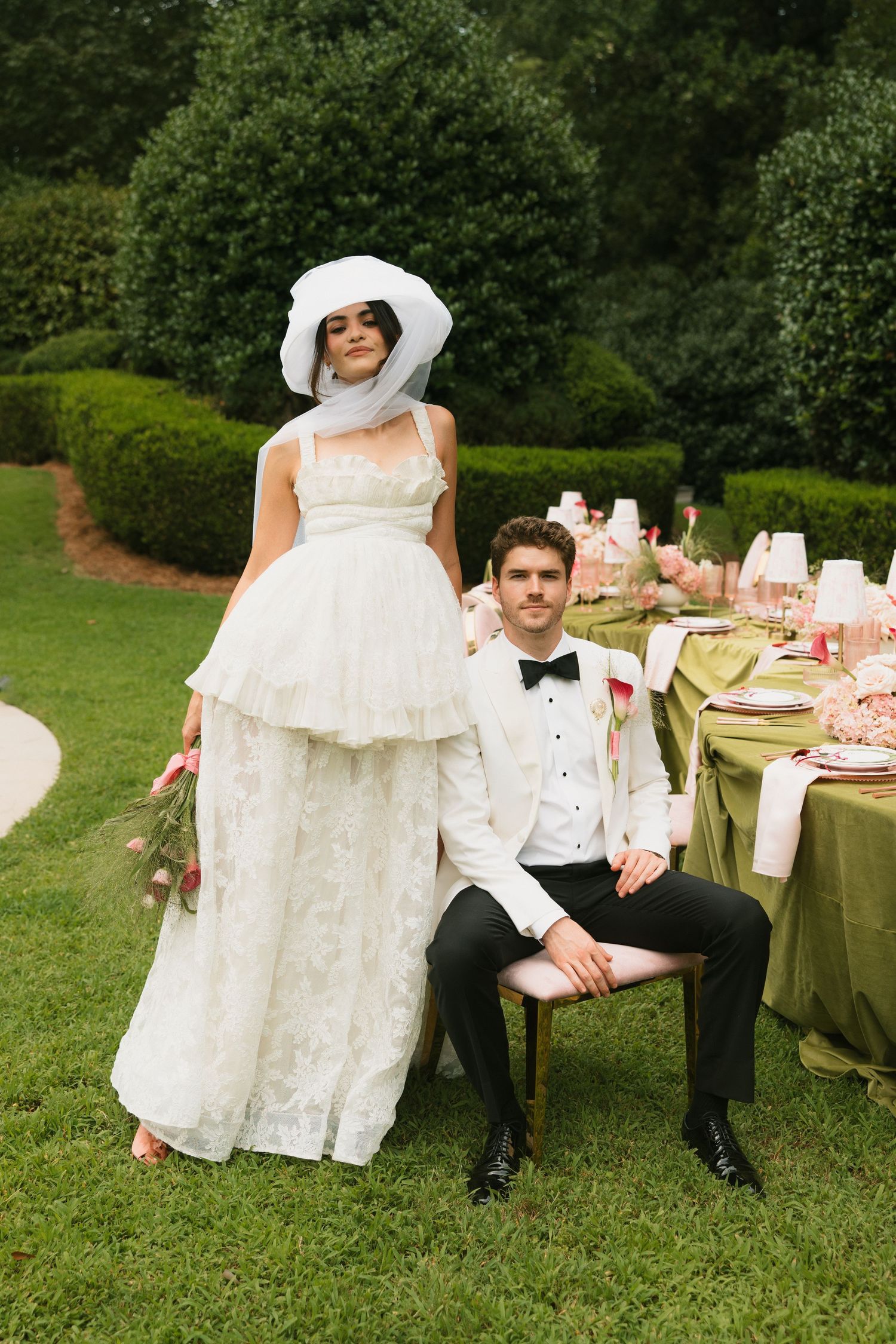 Bride in white gown and hat stands beside groom in tuxedo at elegant outdoor wedding reception table.