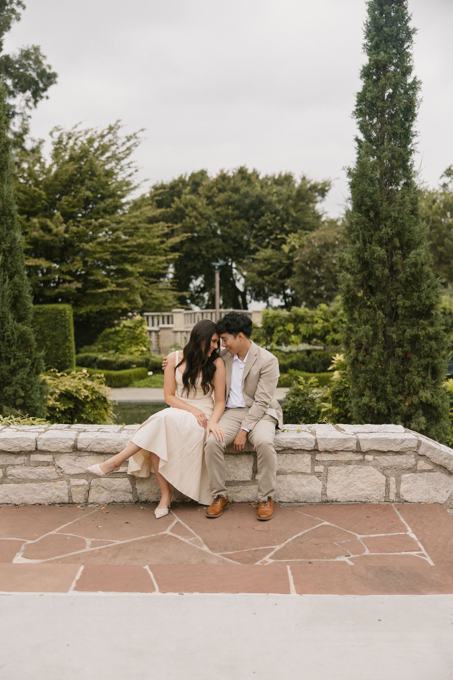 Couple sitting close on stone wall in garden, leaning foreheads together in romantic outdoor setting.