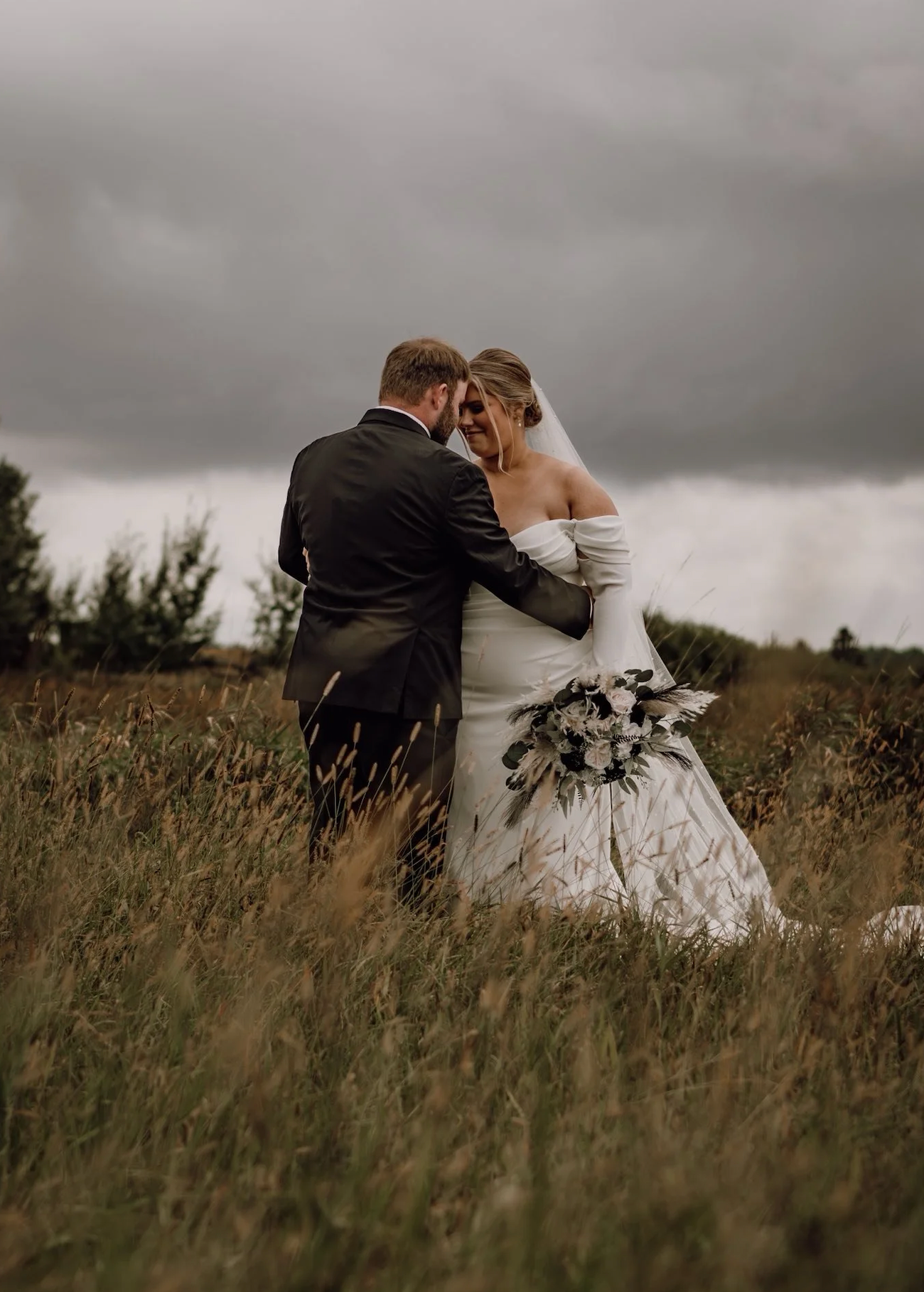 A bride and groom embrace in a misty field, she holds a bouquet in a white gown as storm clouds gather overhead.