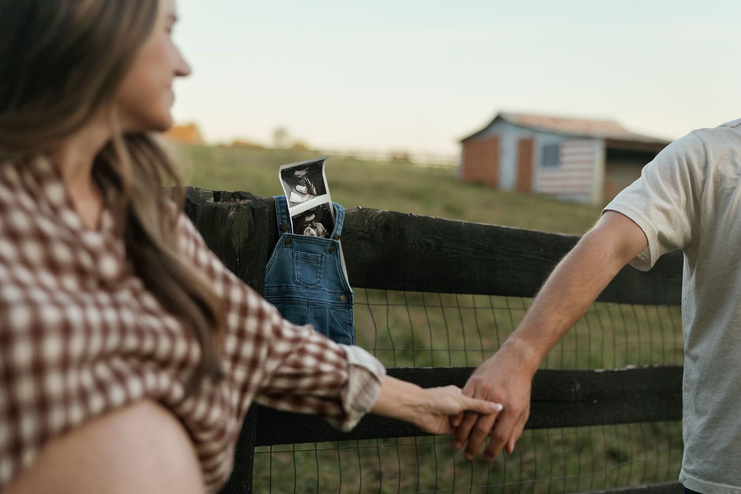Pregnant couple holding hands by a wooden fence, with an ultrasound photo tucked in her jeans pocket on a farm.