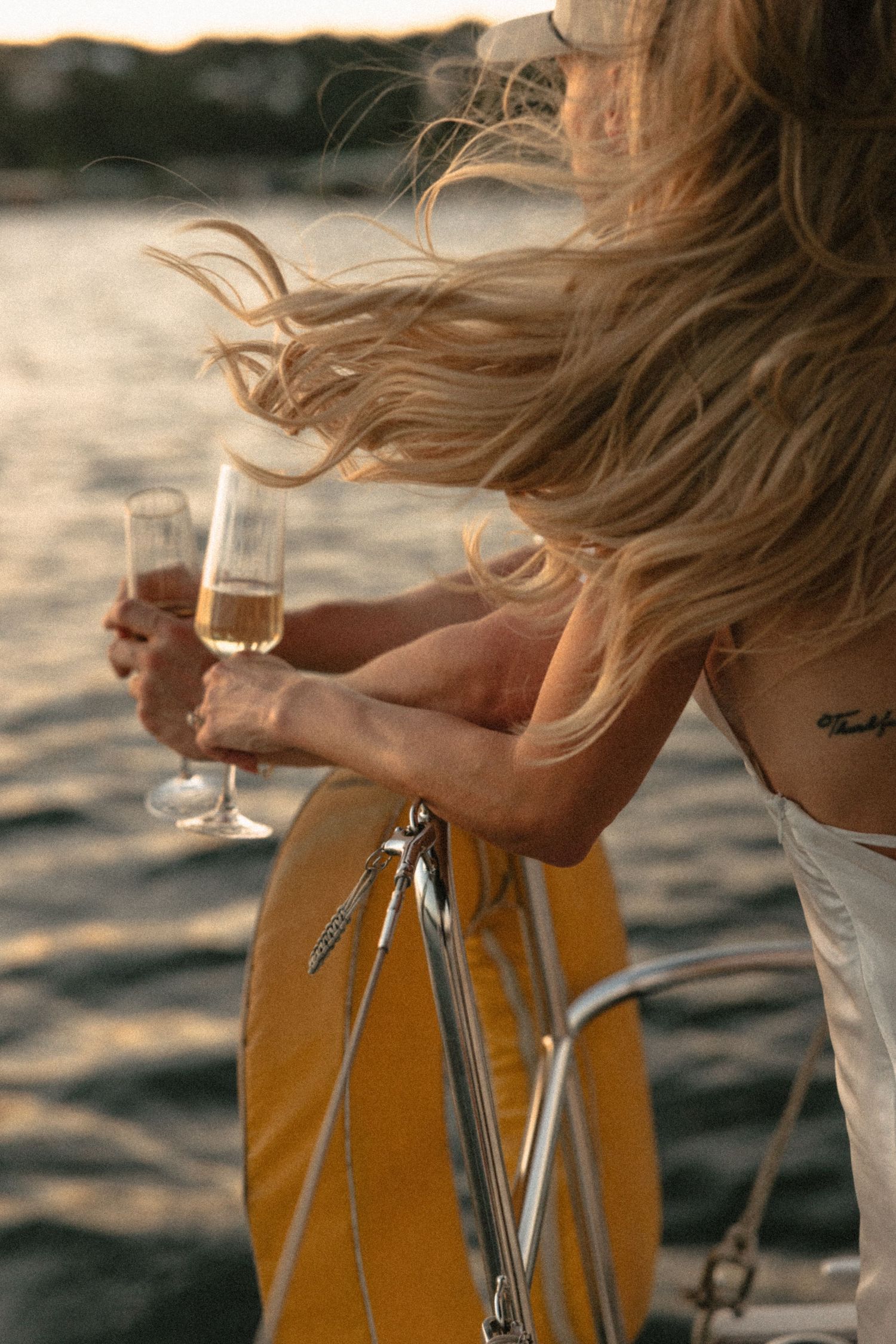 Woman with windswept blonde hair holding champagne glass on sailboat at sunset.