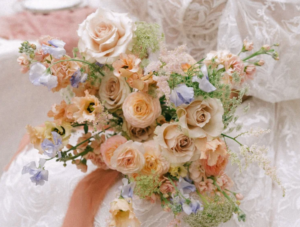 A bridal bouquet featuring peach roses, lavender blooms, and wildflowers held by a bride in a white lace gown.