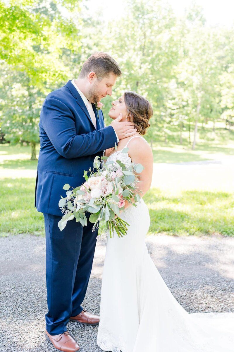 Bride and groom share romantic moment outdoors, groom in navy suit cradling bride's face, bride holding lush pink bouquet.