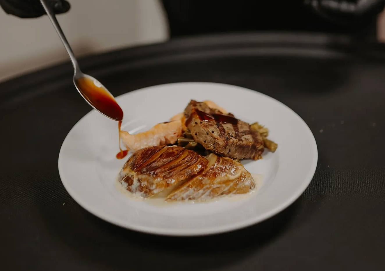 A spoon drizzling rich brown sauce over a gourmet meat dish with caramelized onions on a white plate.