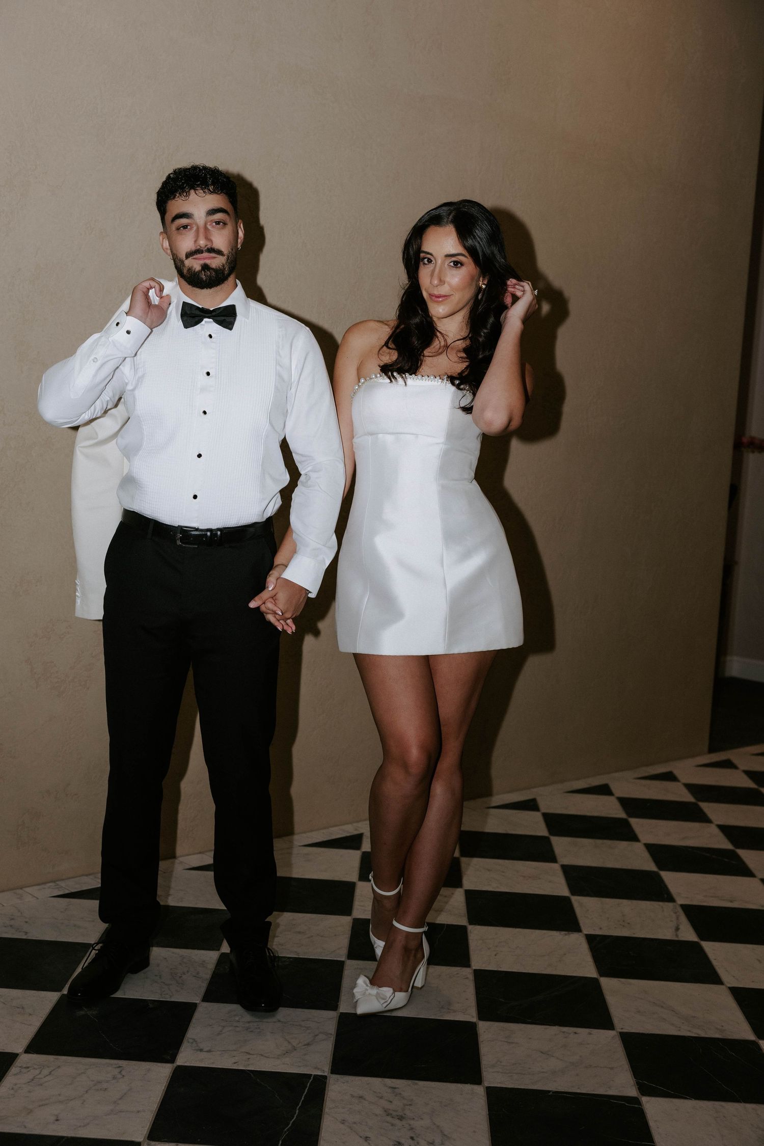 Couple posing on checkered floor, man in tuxedo and woman in white mini dress, holding hands at wedding reception.