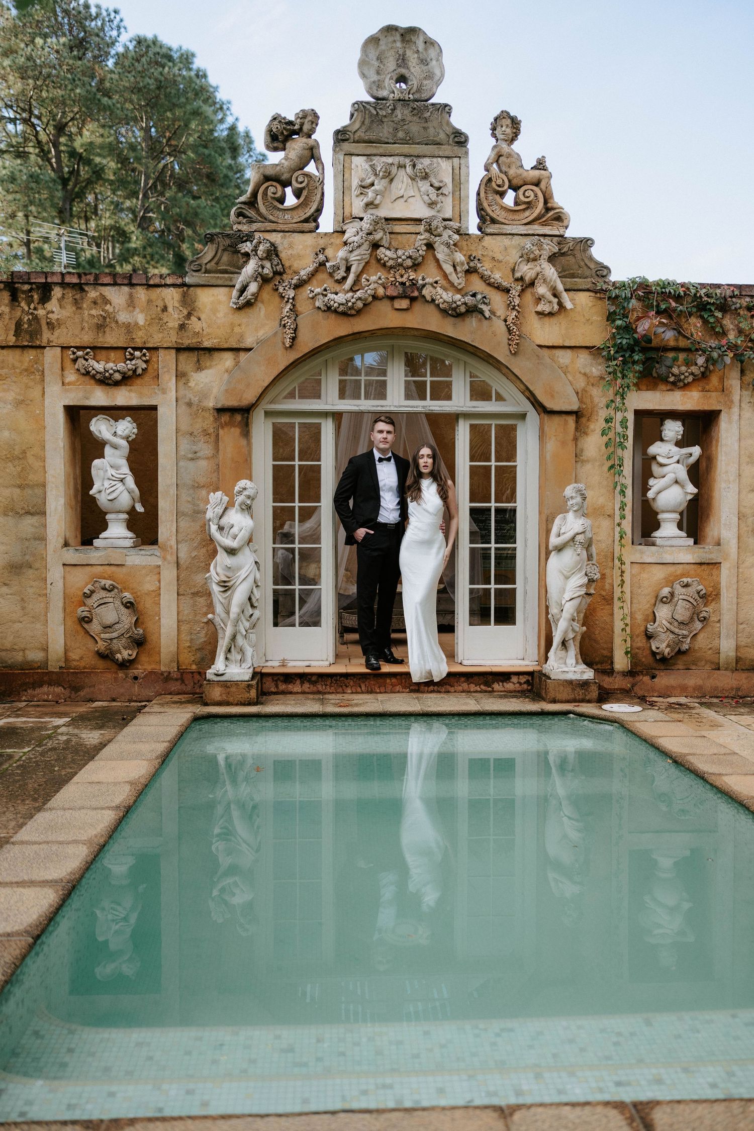 Elegant wedding couple poses by ornate baroque pool with classical statues and French doors at luxury venue.