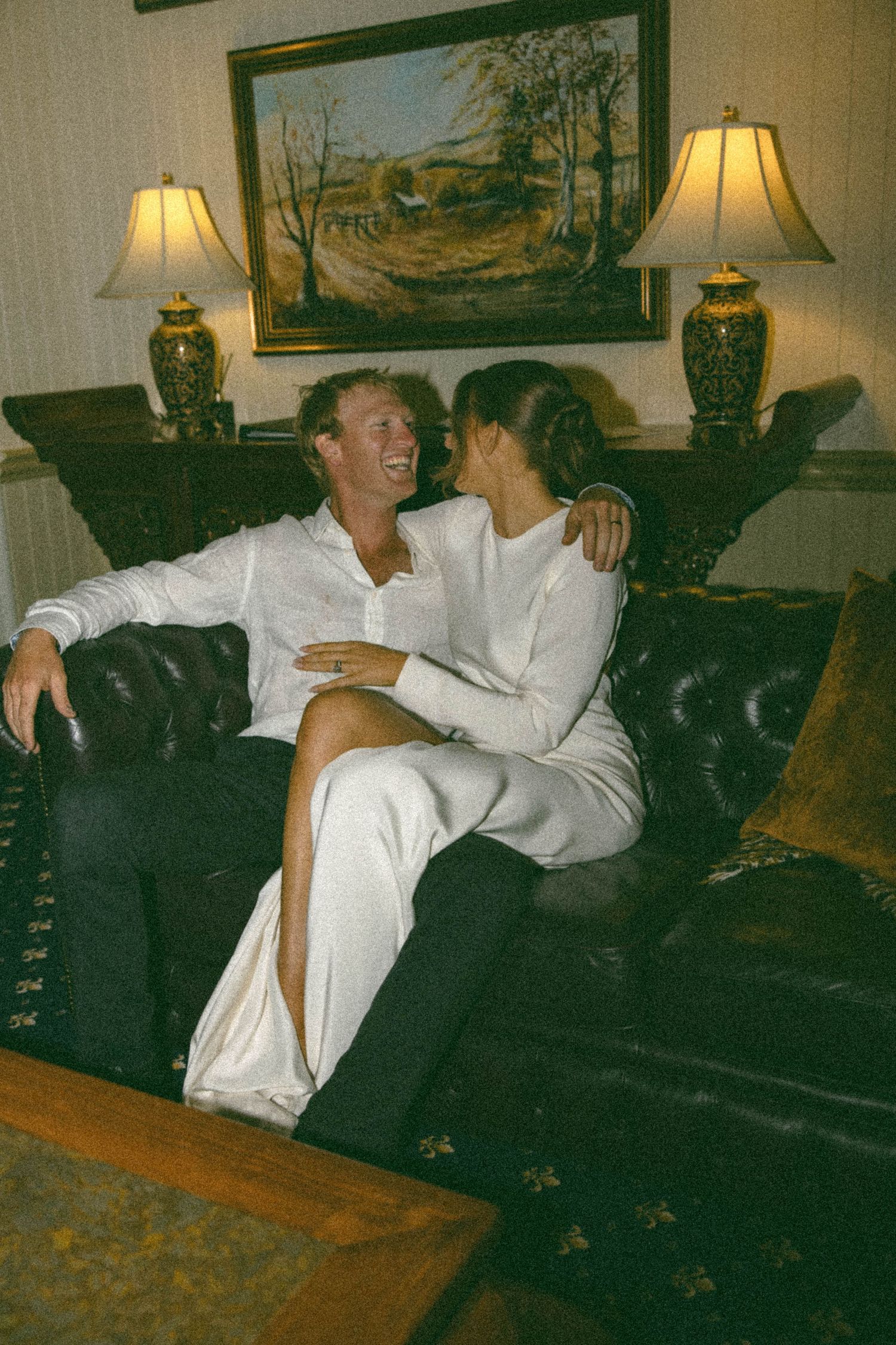 Couple in white wedding attire laughing together on a leather sofa with lamps and painting backdrop.