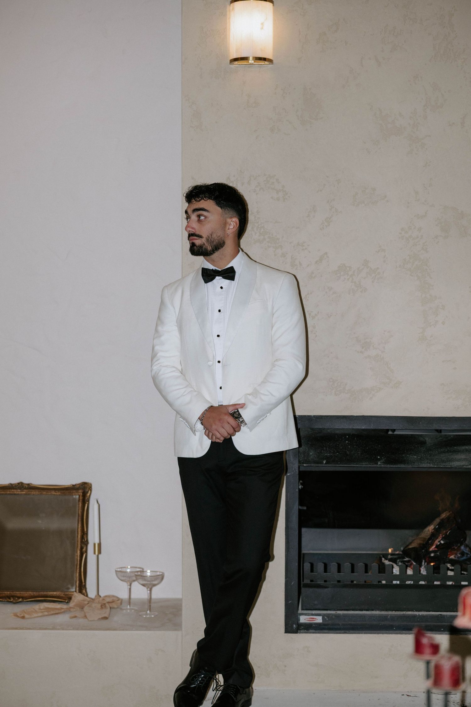 A man in a white tuxedo jacket and black bow tie stands elegantly by a fireplace in a formal indoor setting.