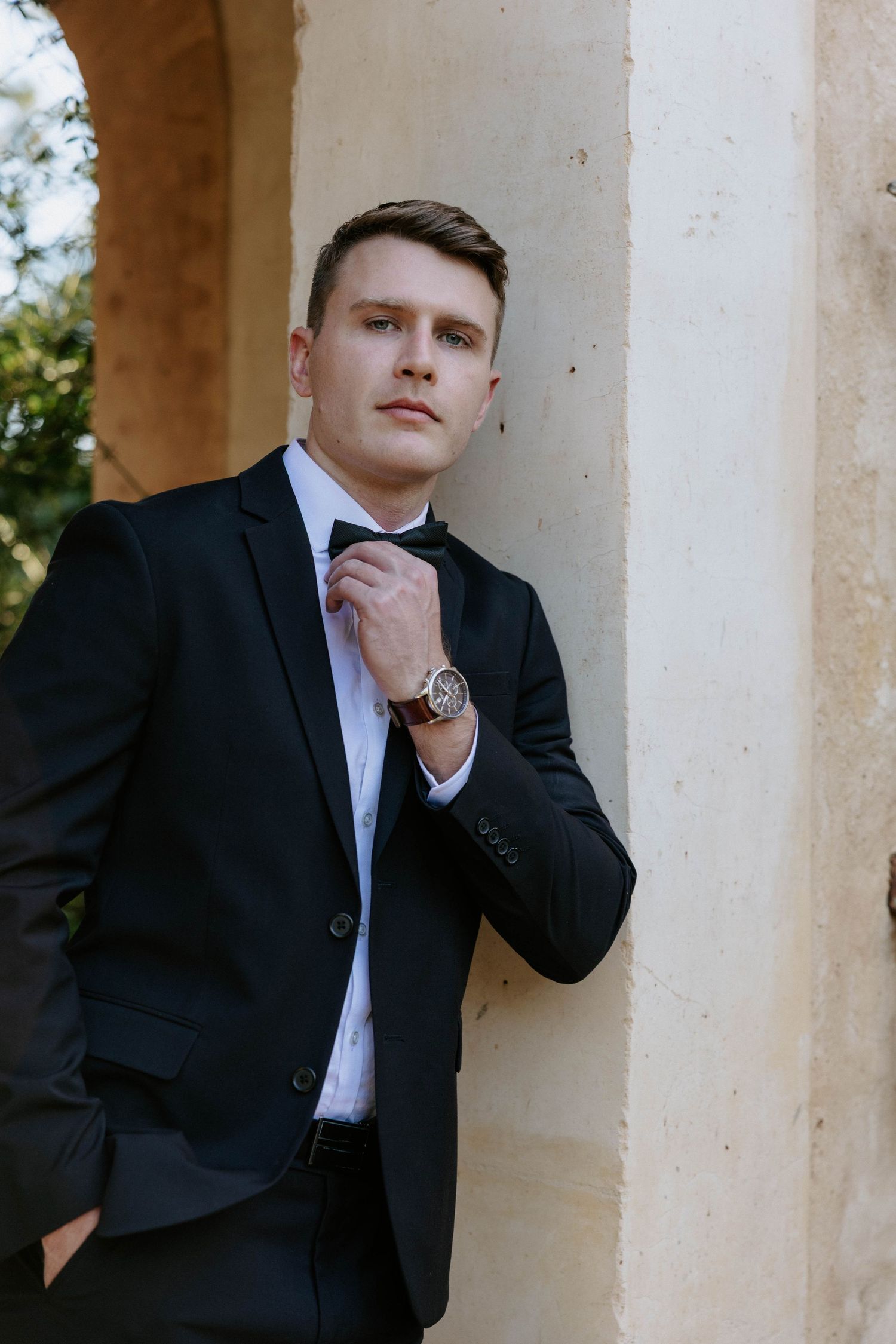 Handsome groom in black tuxedo adjusting bow tie while leaning against a stone column outdoors.