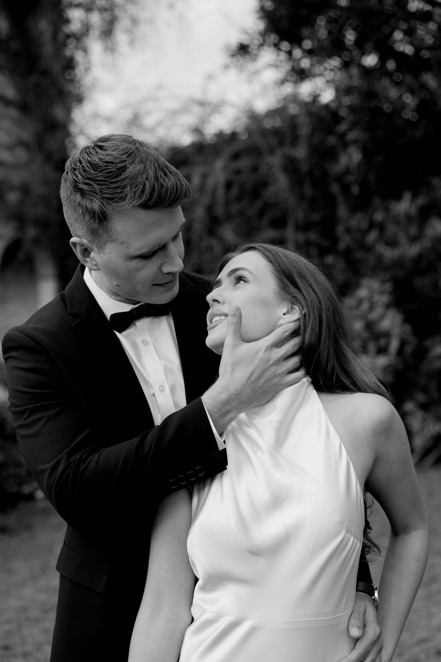 Black and white photo of a groom in a tuxedo tenderly holding his bride's face as they gaze at each other outdoors.